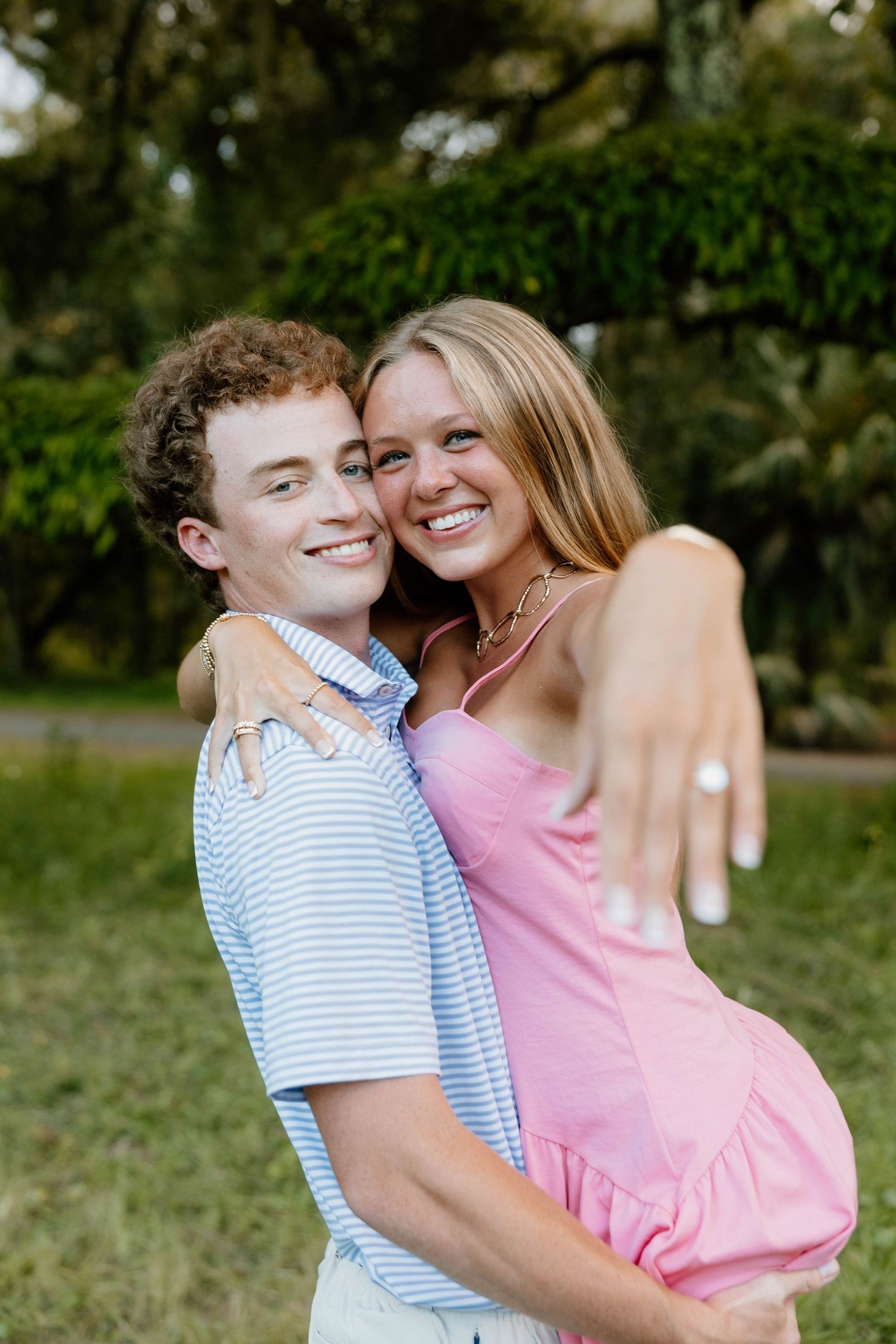Happy young couple embracing outdoors, woman in pink dress showing engagement ring, smiling in lush green park setting.