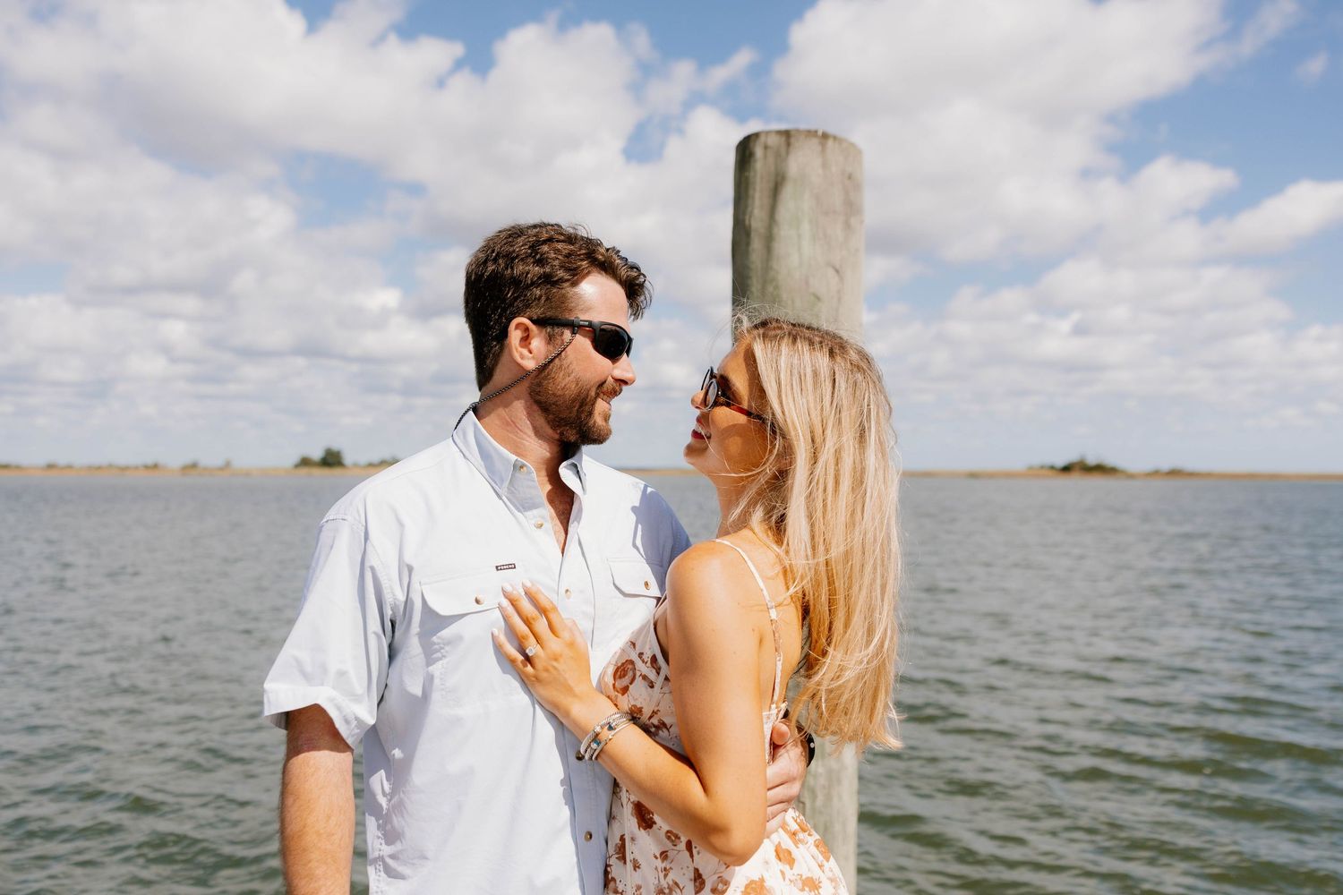 Happy couple smiling at each other near a wooden pier post with scenic water background.