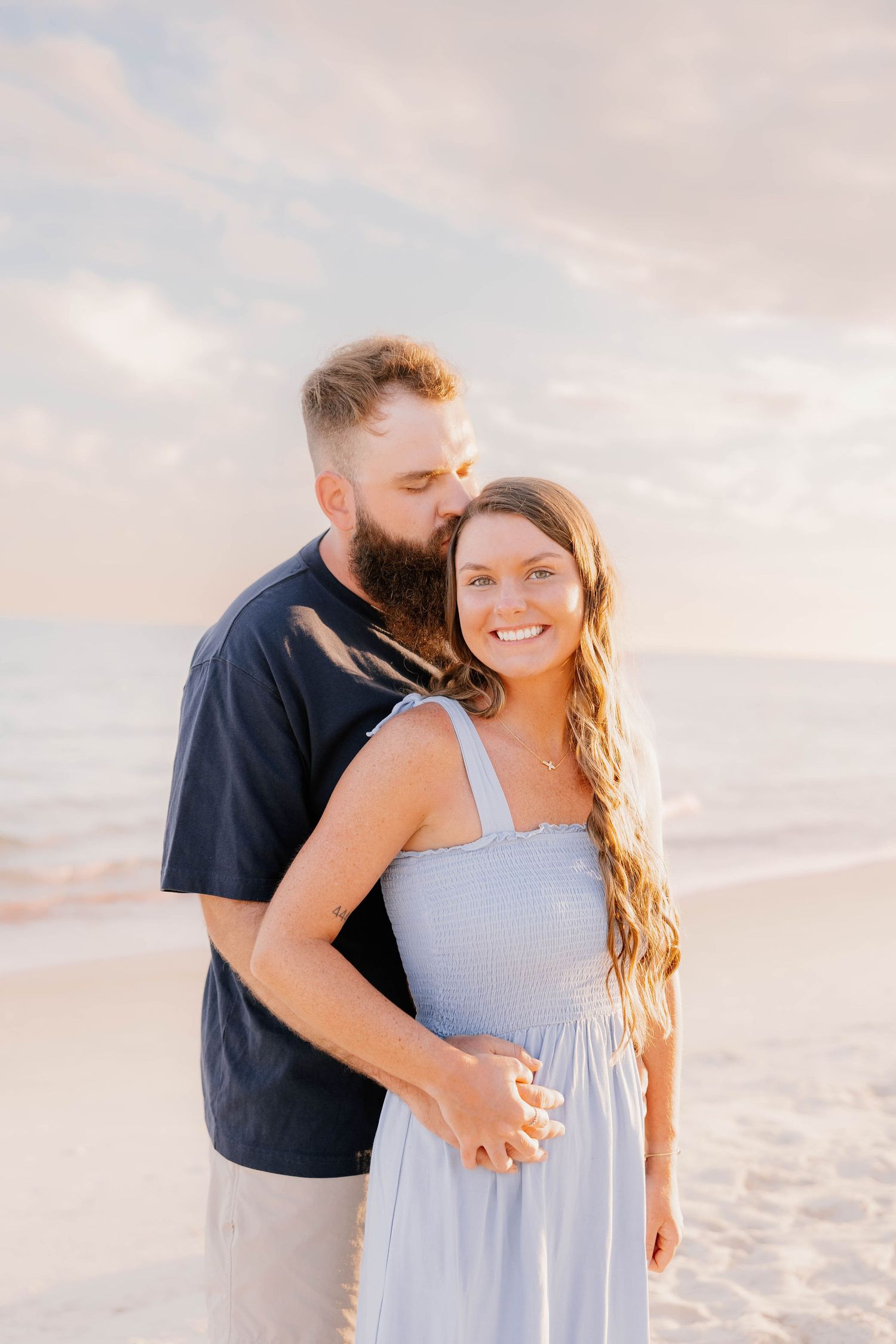 A couple embraces on a beach at sunset, the man kissing the womans head as she smiles in a light blue dress.
