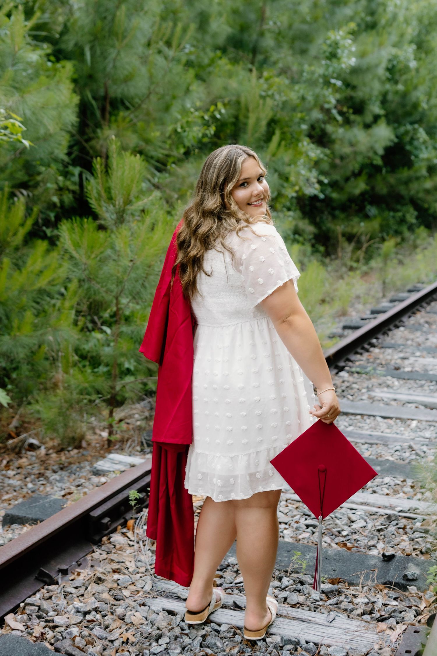 Smiling graduate in white dress holds red cap and gown on railroad tracks surrounded by lush green trees.
