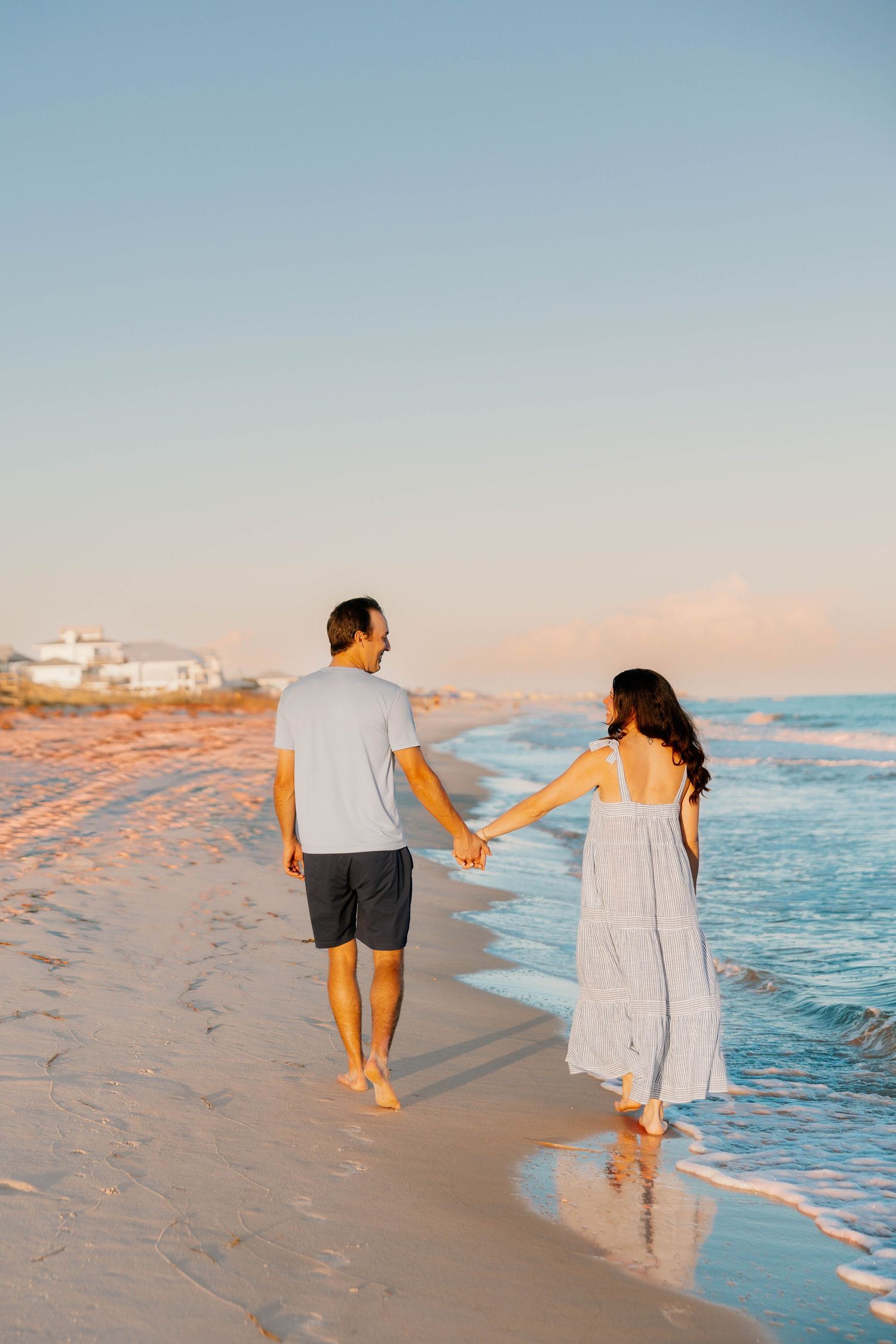 Couple holding hands walking along a sandy beach at sunset, man in gray shirt and woman in flowing white dress.