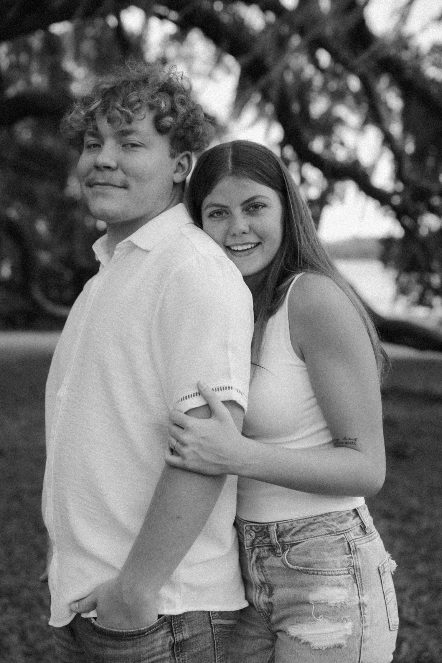 A black and white portrait of a couple in casual attire smiling together outdoors.