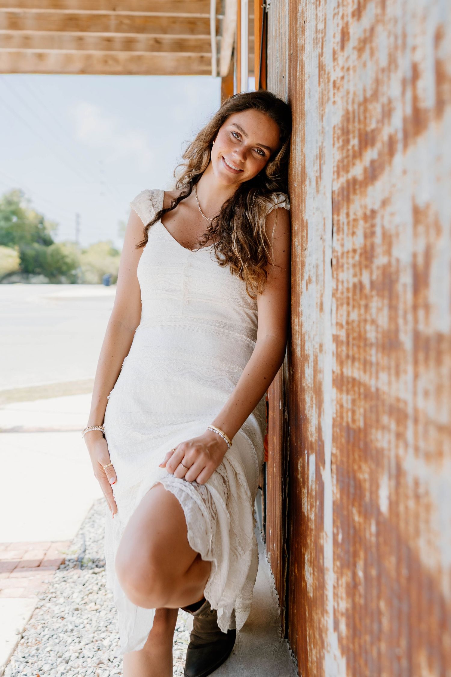 Young woman in white lace dress leaning against rustic wall, smiling outdoors in natural light.