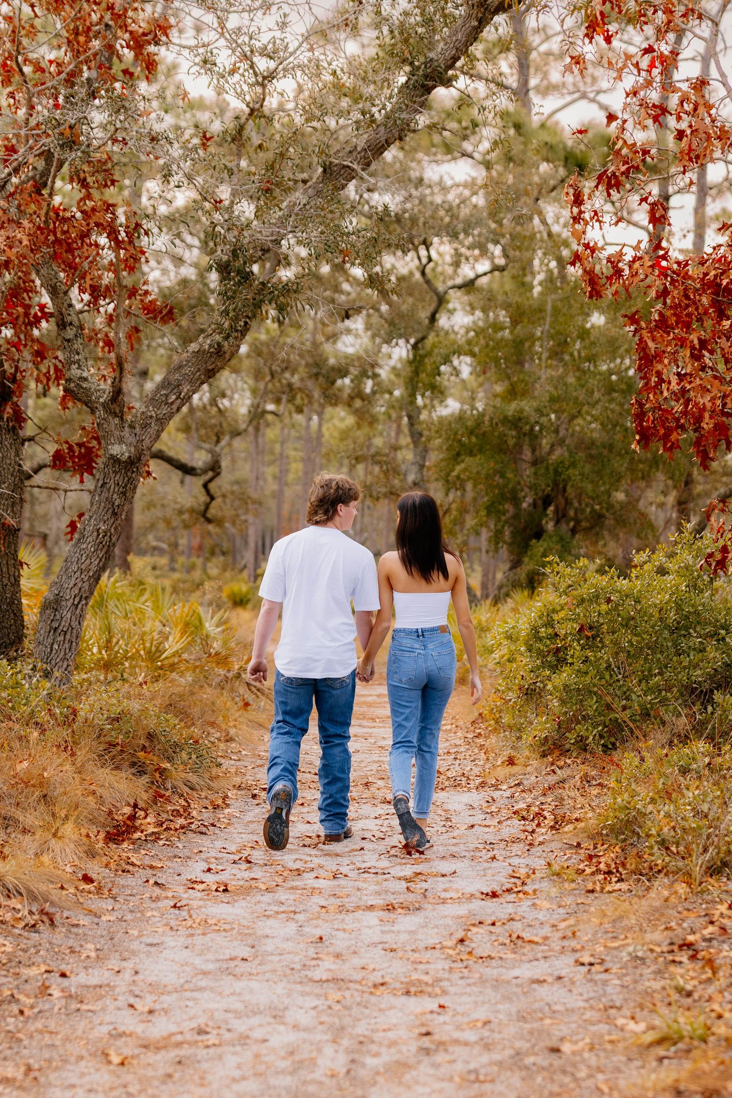 Couple holding hands walking down a scenic autumn forest path surrounded by red foliage and tall trees.