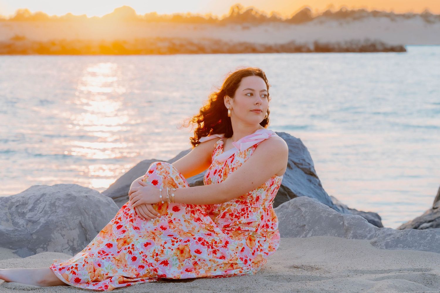 Woman in floral dress sitting on rocks at golden hour sunset by the ocean.
