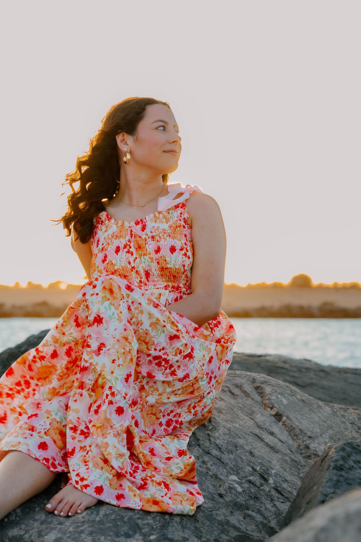 Woman in floral dress sitting on rocks by water at golden hour sunset.