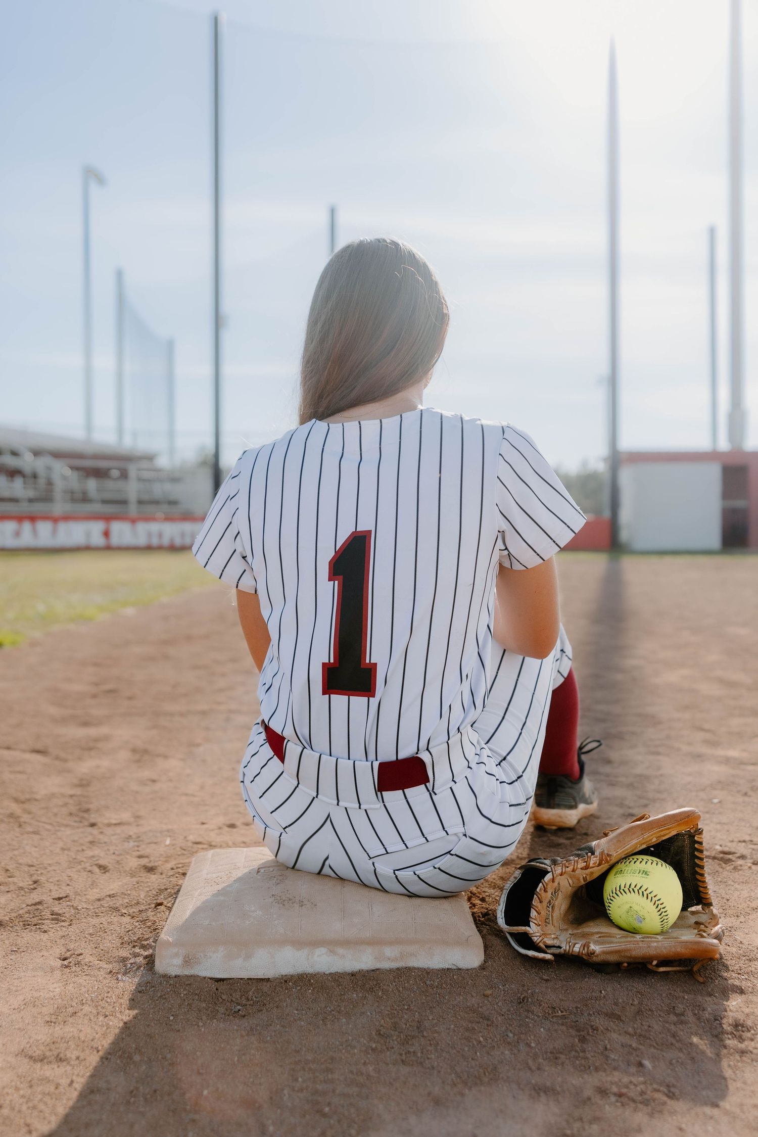 Softball player in pinstripe jersey number 1 sits on base with glove and ball on a dirt field.