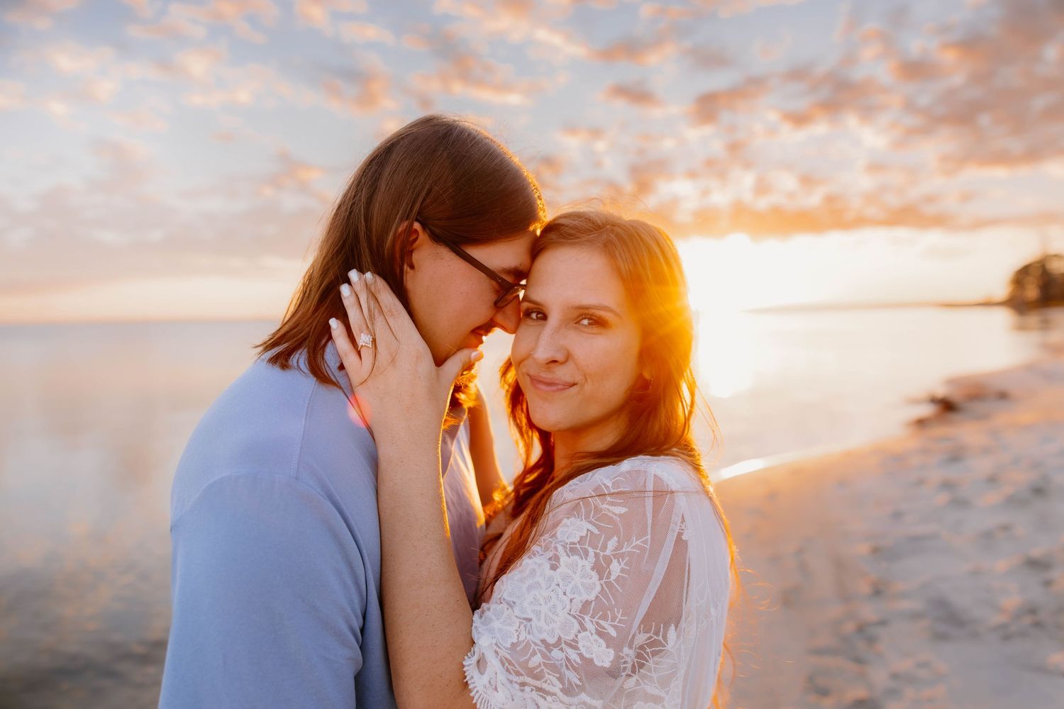 Couple sharing a romantic sunset moment on a beach, woman smiling at camera in a white lace top.