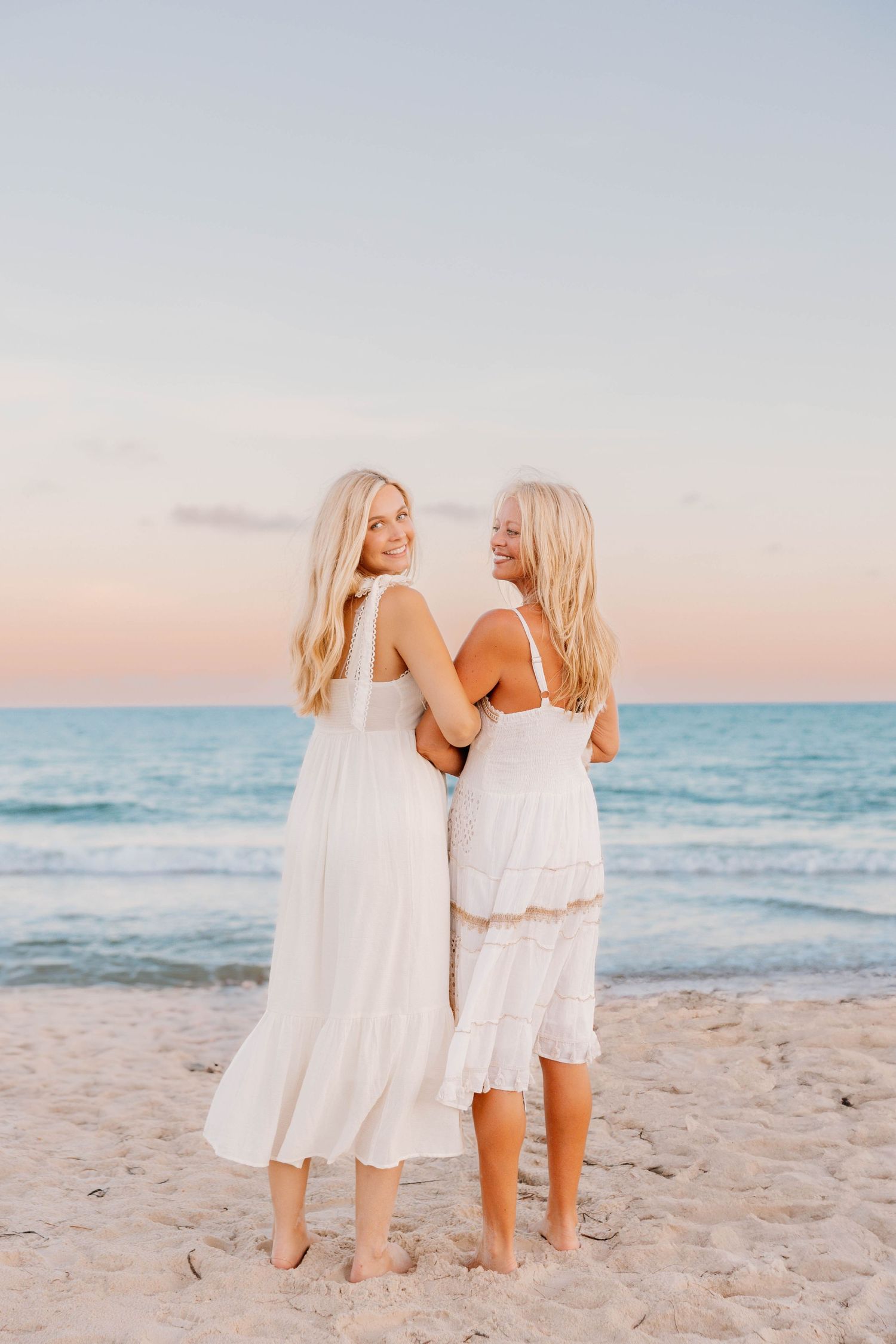 Two children in white dresses walking together on a sandy beach at sunset with ocean waves in the background.