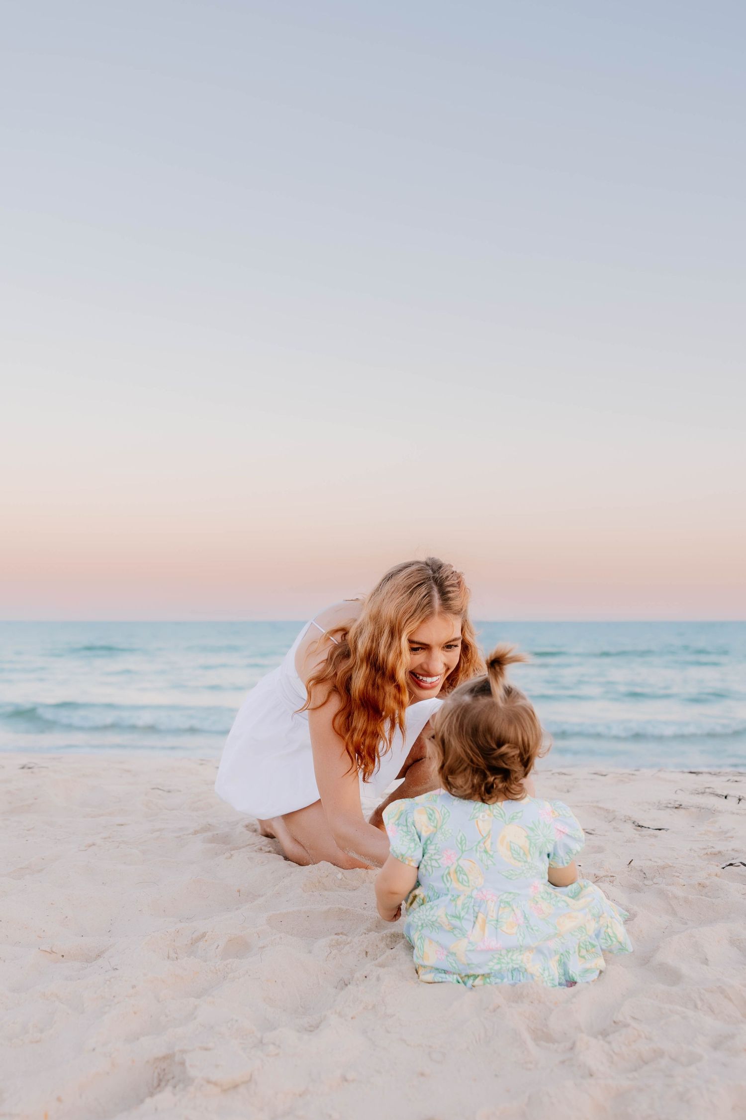 A smiling mother in a white dress plays with her baby on a sandy beach at sunset.