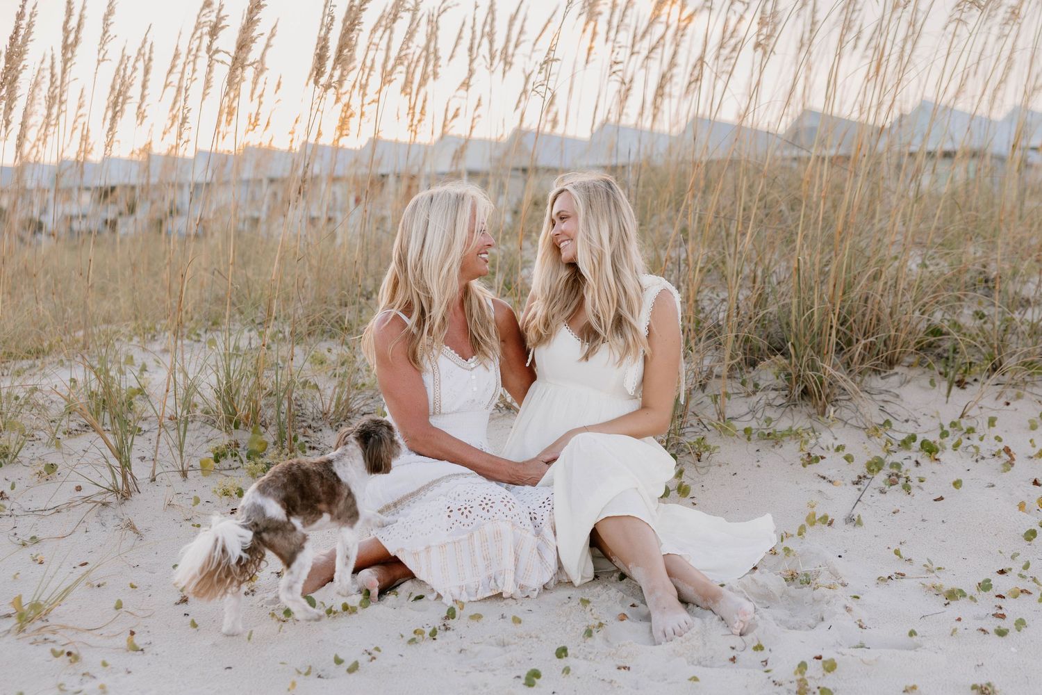 Two people in white dresses sit together on a sandy beach with tall grass and mountains in the background.