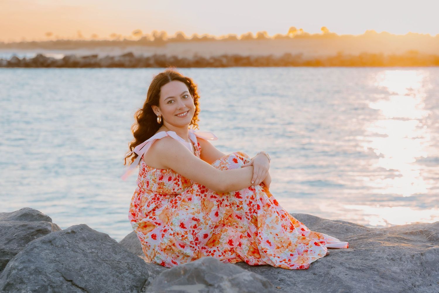Woman in floral dress sitting on rocks by the water at golden hour sunset, smiling over shoulder.