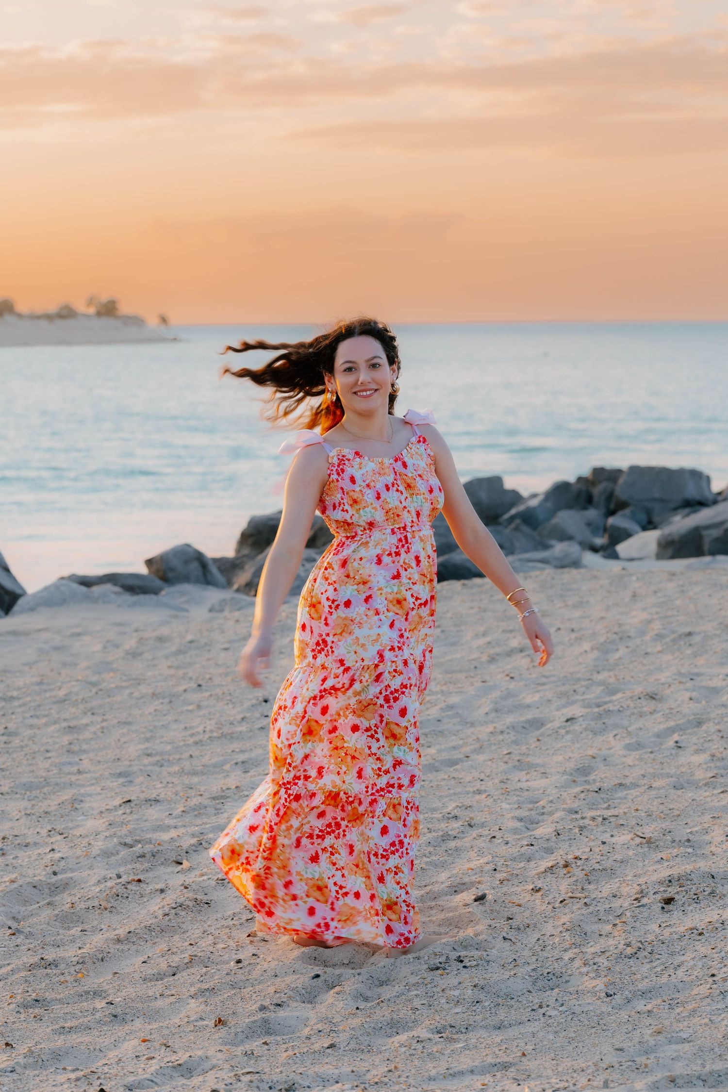 Woman in floral maxi dress walking on sandy beach at golden sunset near rocky shore.
