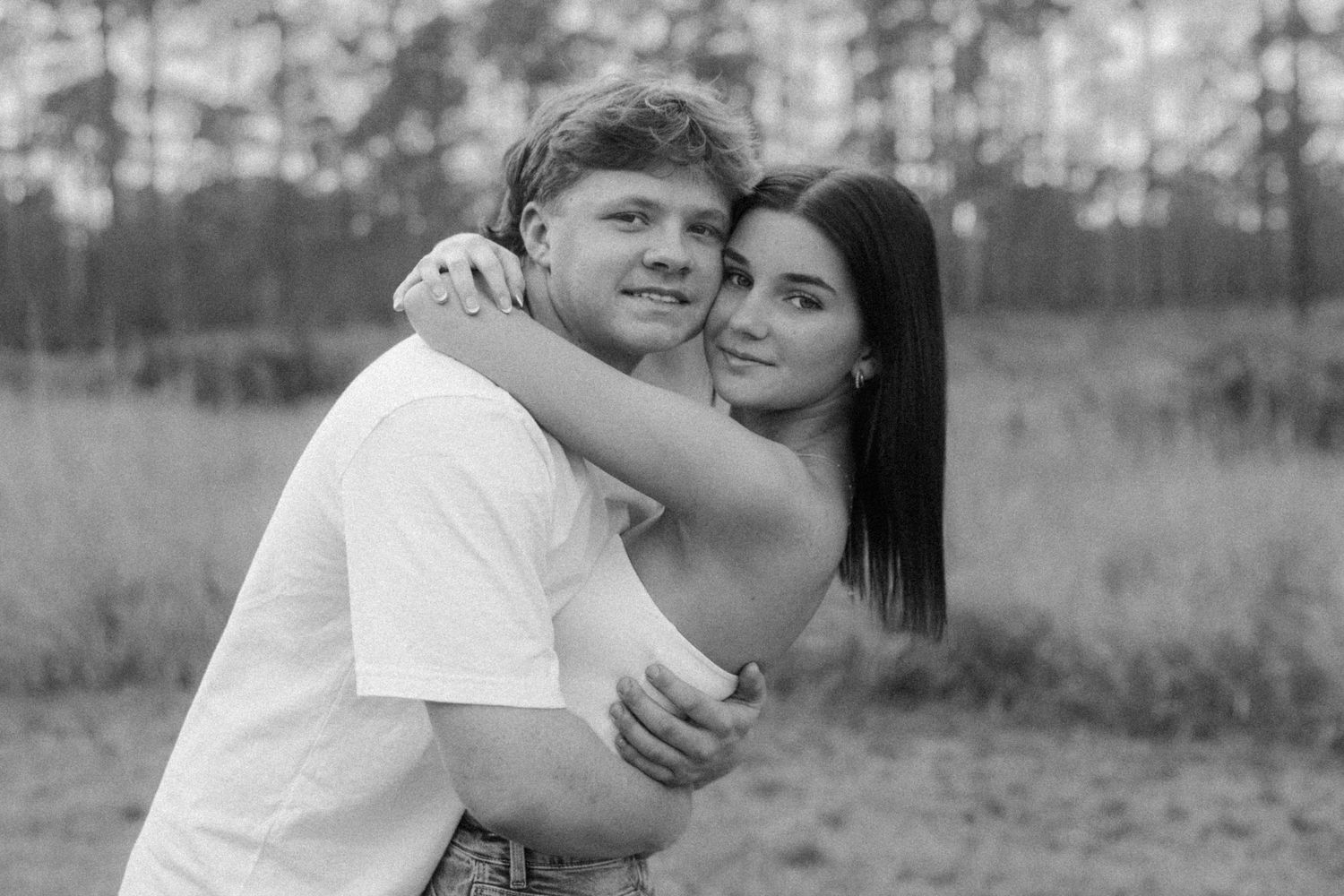 Black and white photo of a young couple embracing outdoors, woman hugging man from behind, pine trees in background.