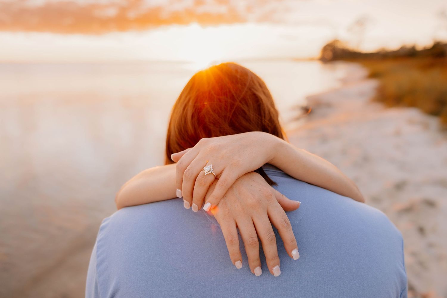 Close-up of woman hugging partner at beach sunset, showcasing diamond engagement ring on her hand.