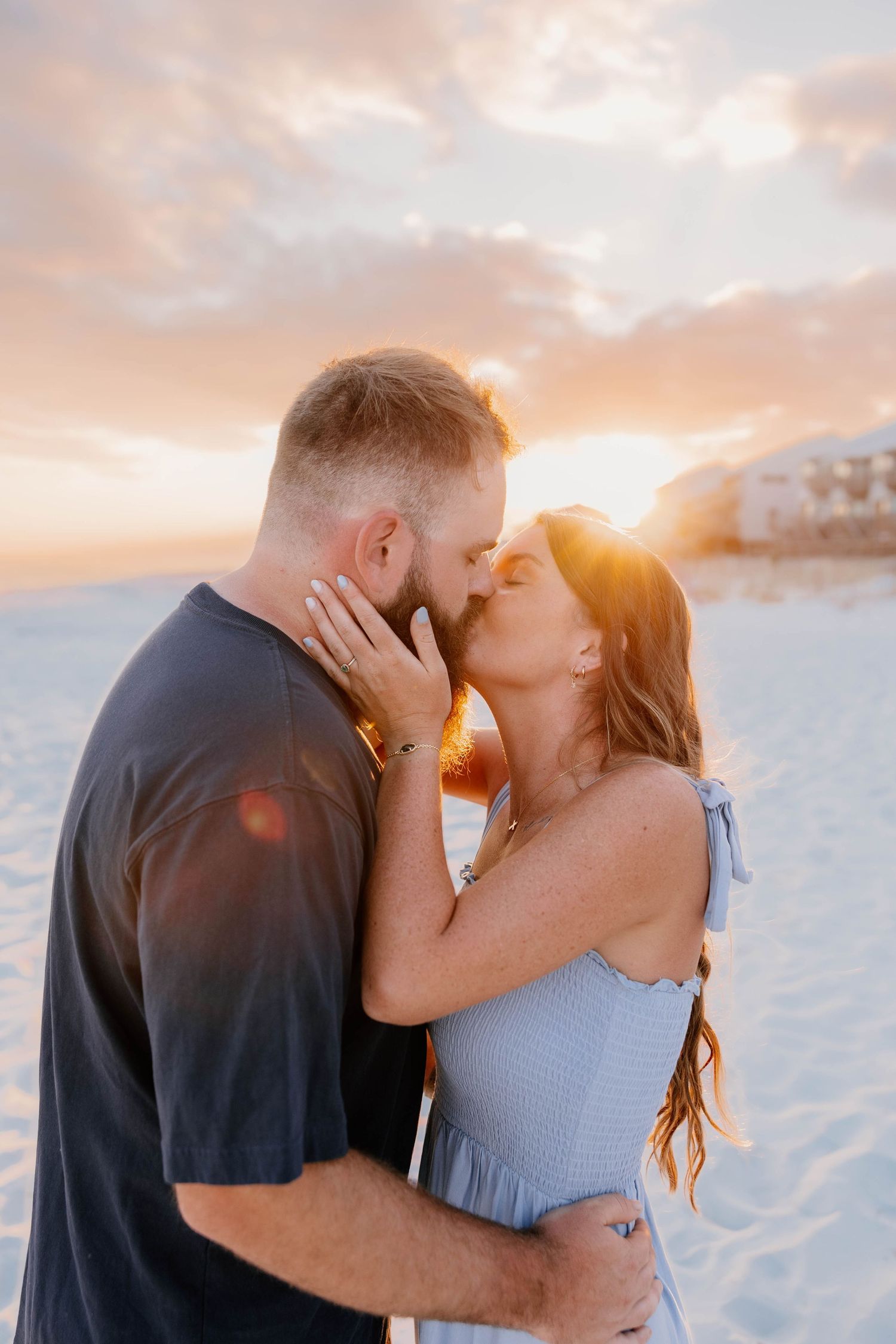 Couple sharing a romantic kiss on a sandy beach at golden sunset, woman in blue dress tenderly touching mans face.