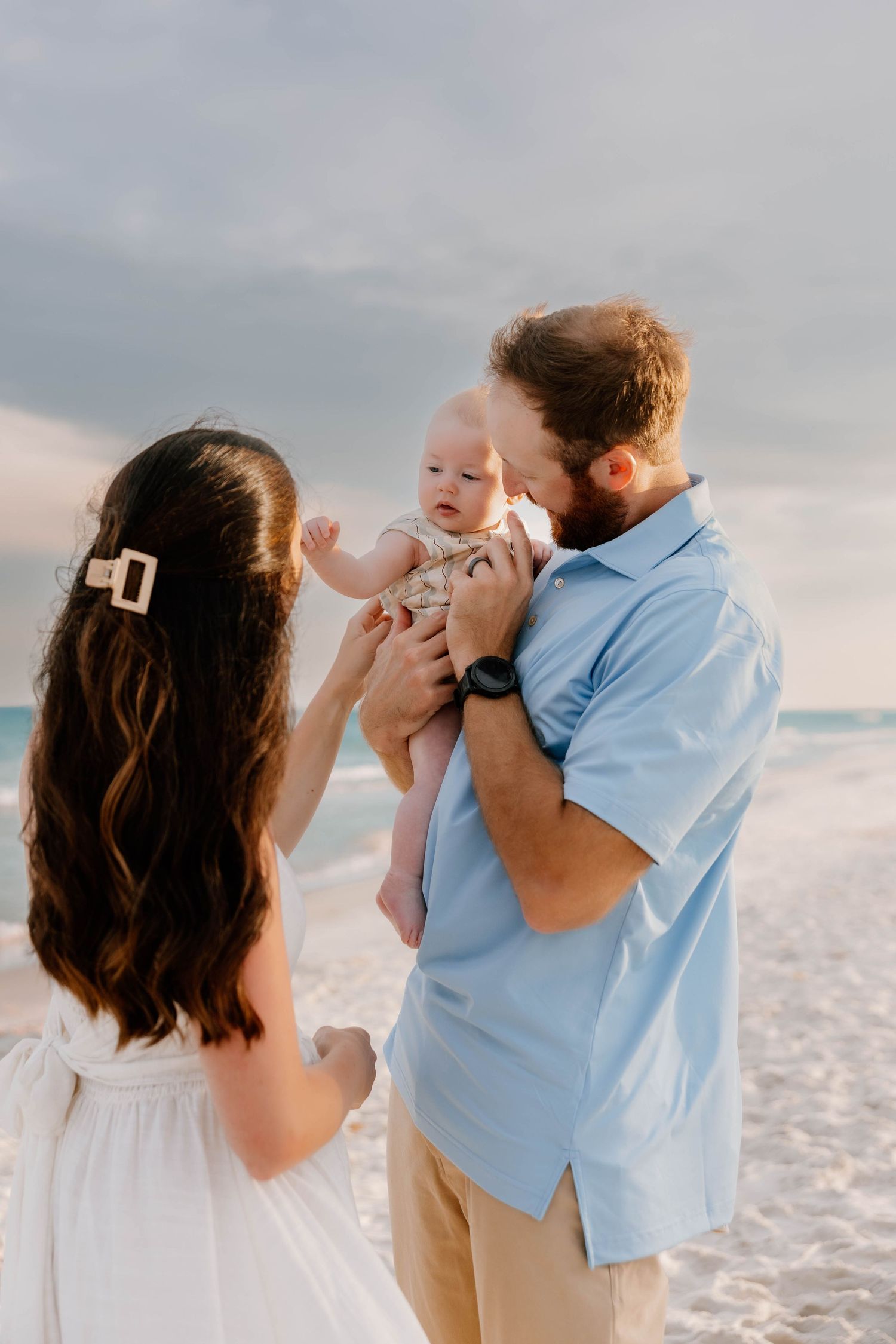 A couple shares a tender moment with their baby on a beach, the father kissing the infant while the mother looks on lovingly.