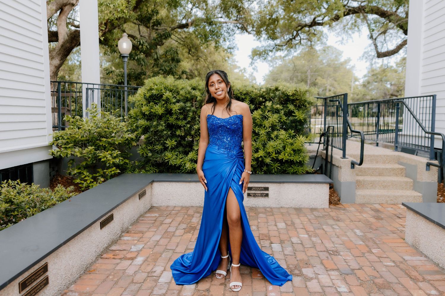 Young woman in royal blue satin halter prom gown with slit poses on brick courtyard outdoors.