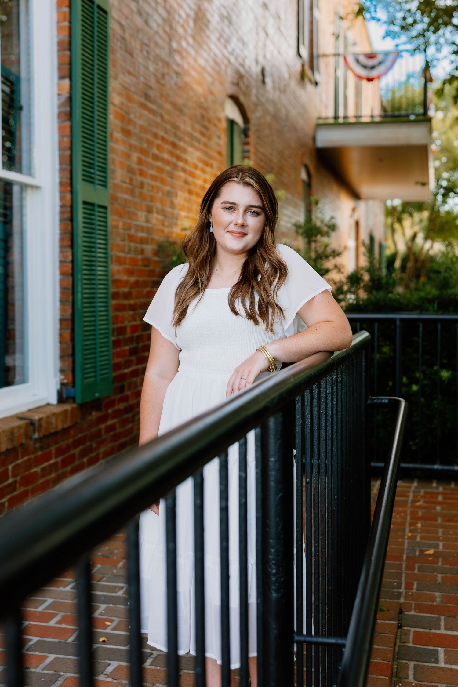 Young woman in white dress posing by black iron railing outside historic brick building with green shutters.