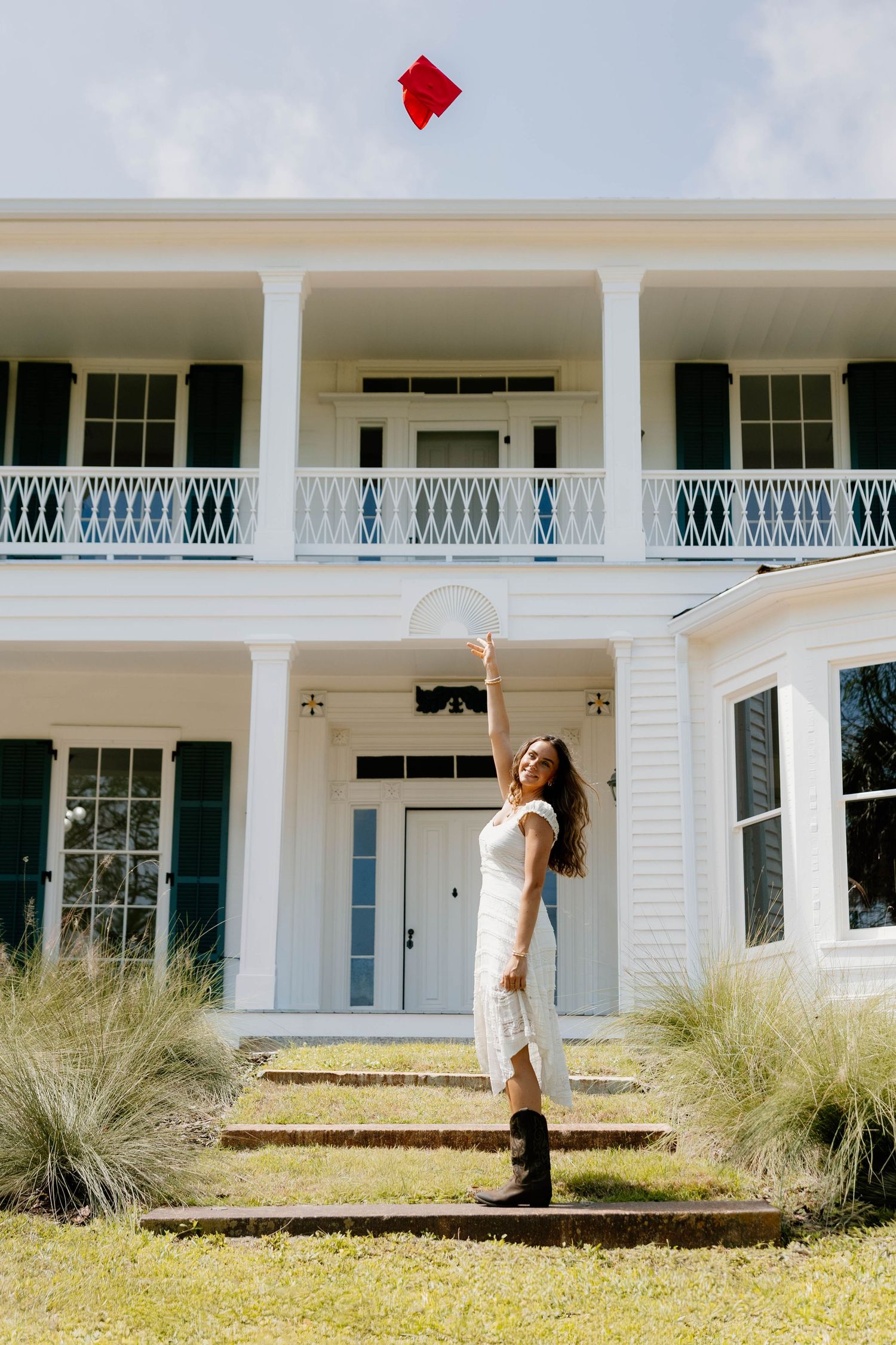 Young woman in white dress and boots tossing red graduation cap in front of elegant white Southern mansion.