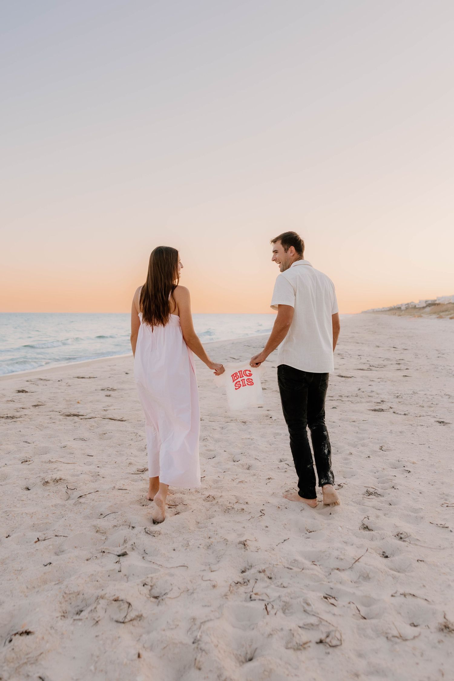 A couple holds hands on a sandy beach at sunset, the woman in a white dress and man in white shirt holding a balloon.