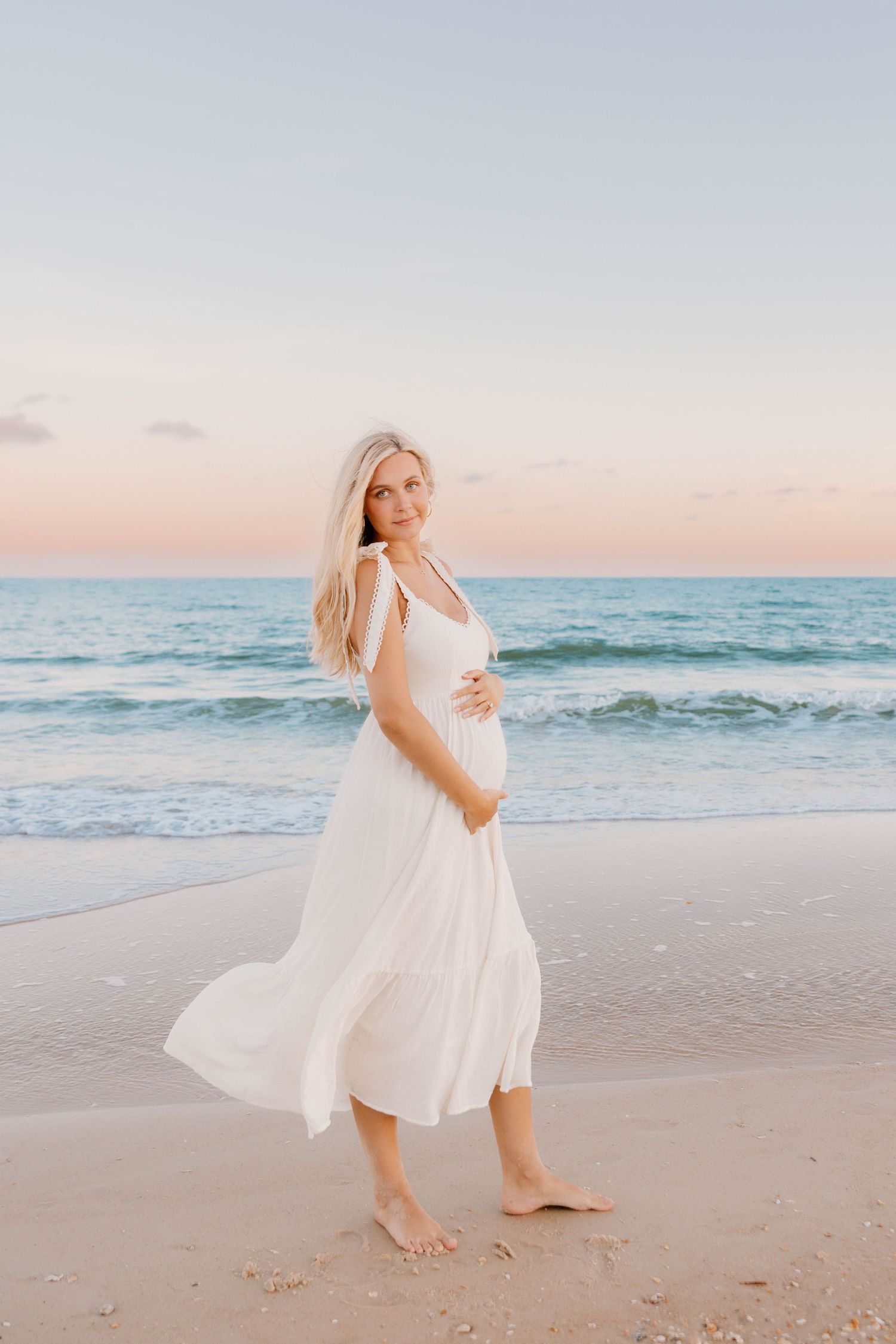 Pregnant woman in flowing white dress walking barefoot on sandy beach at sunset, gently holding her baby bump.