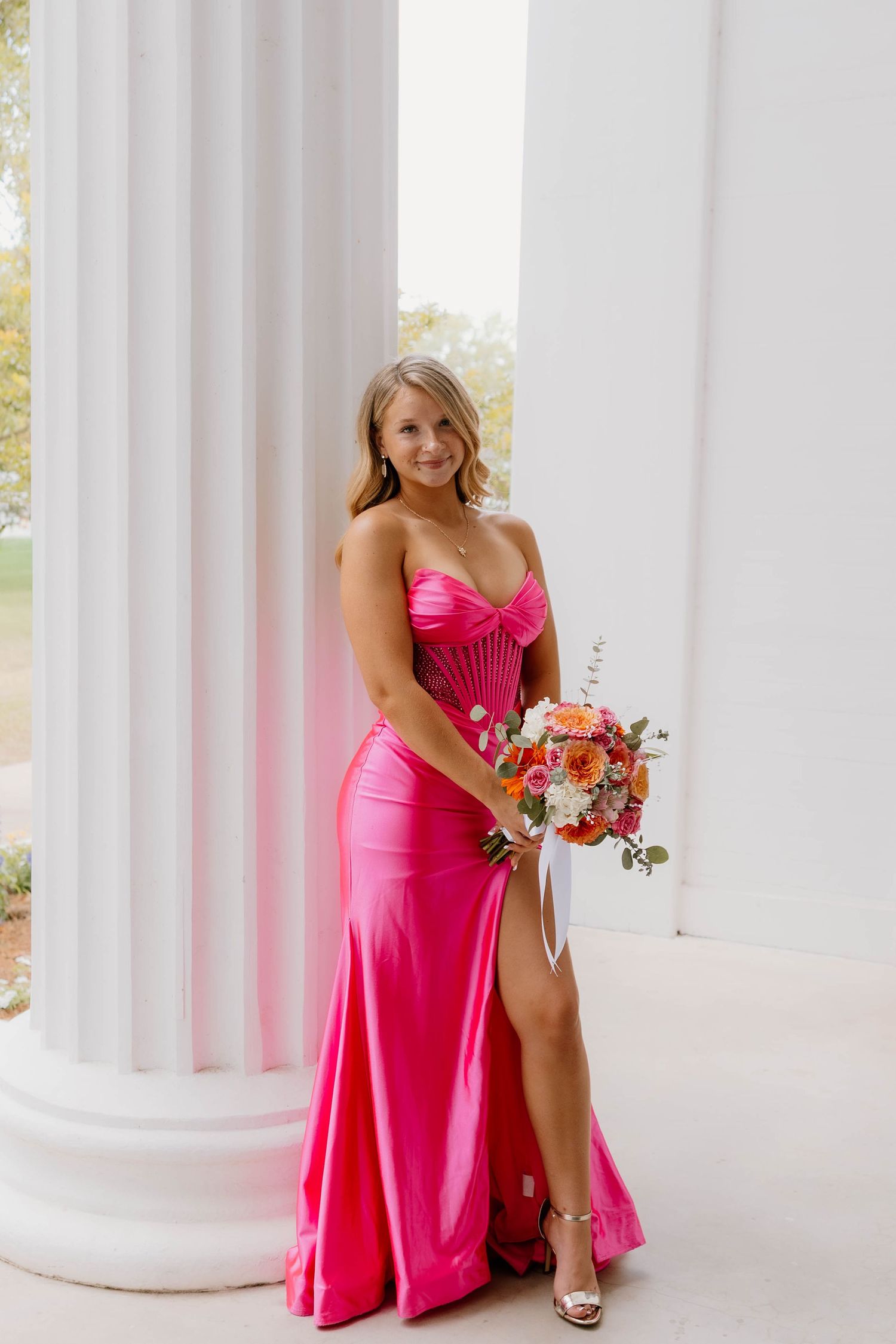 Woman in hot pink strapless satin gown holding colorful bouquet, posing between white columns.