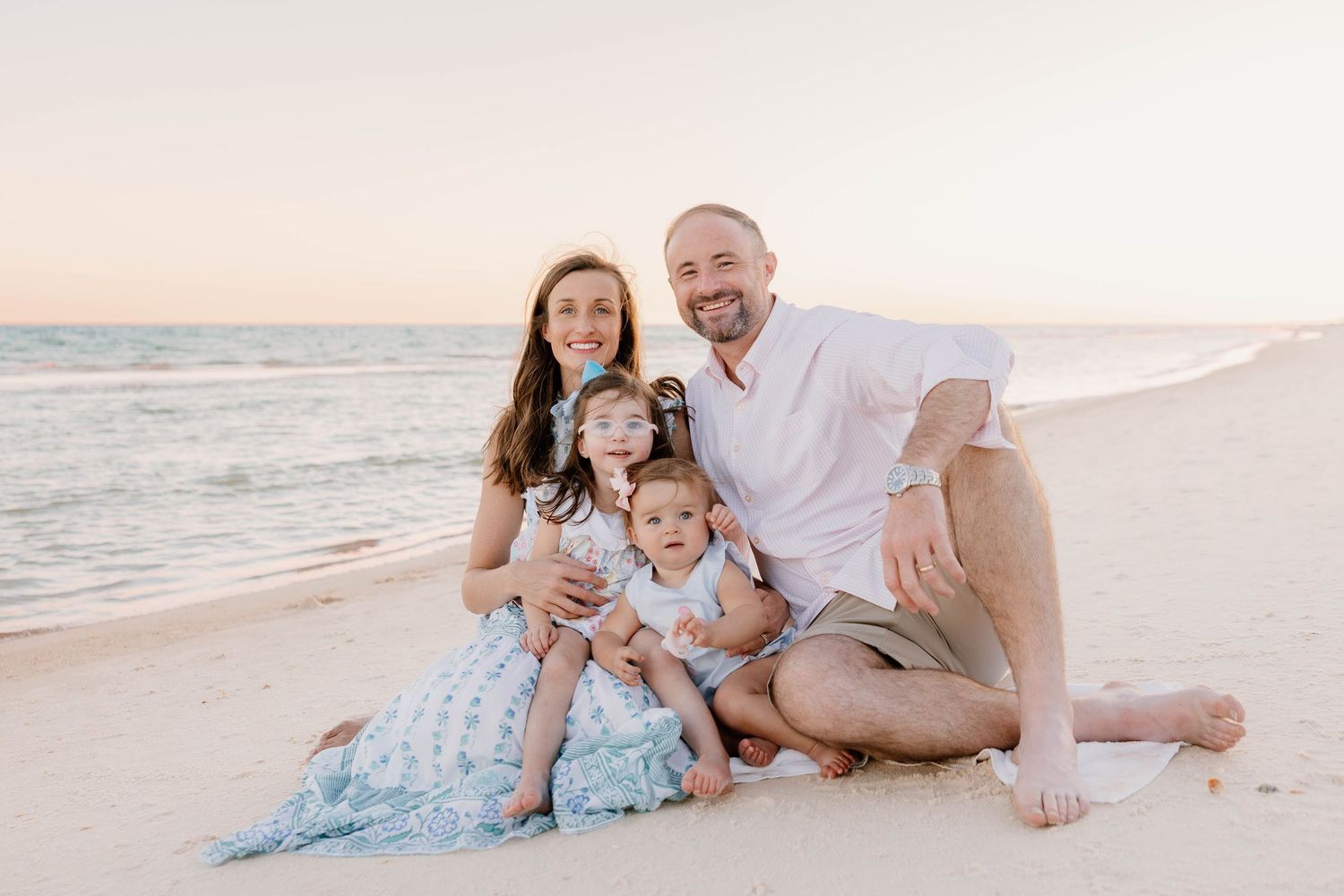 Family of four smiling together on a sandy beach at sunset, parents with two young children in a warm, golden light.