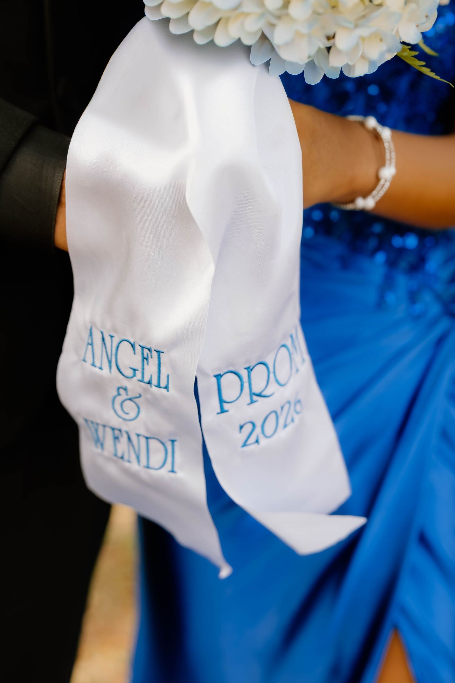 White satin prom corsage ribbon embroidered with Angel & Wendi Prom 2026 in blue lettering on a blue dress.