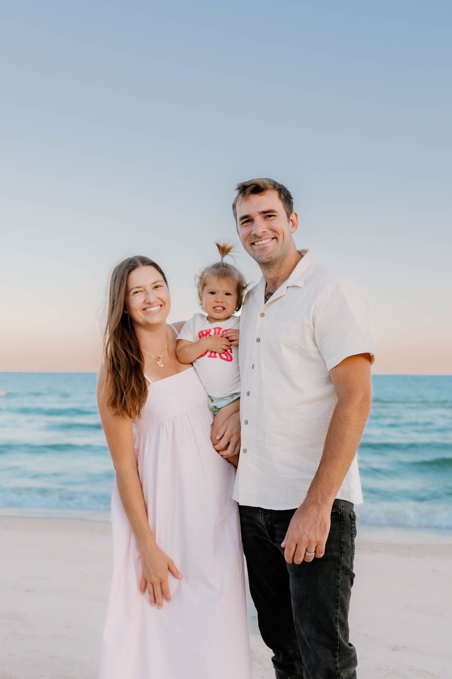 Family enjoying sunset together at beach with ocean waves and soft pastel sky in background.