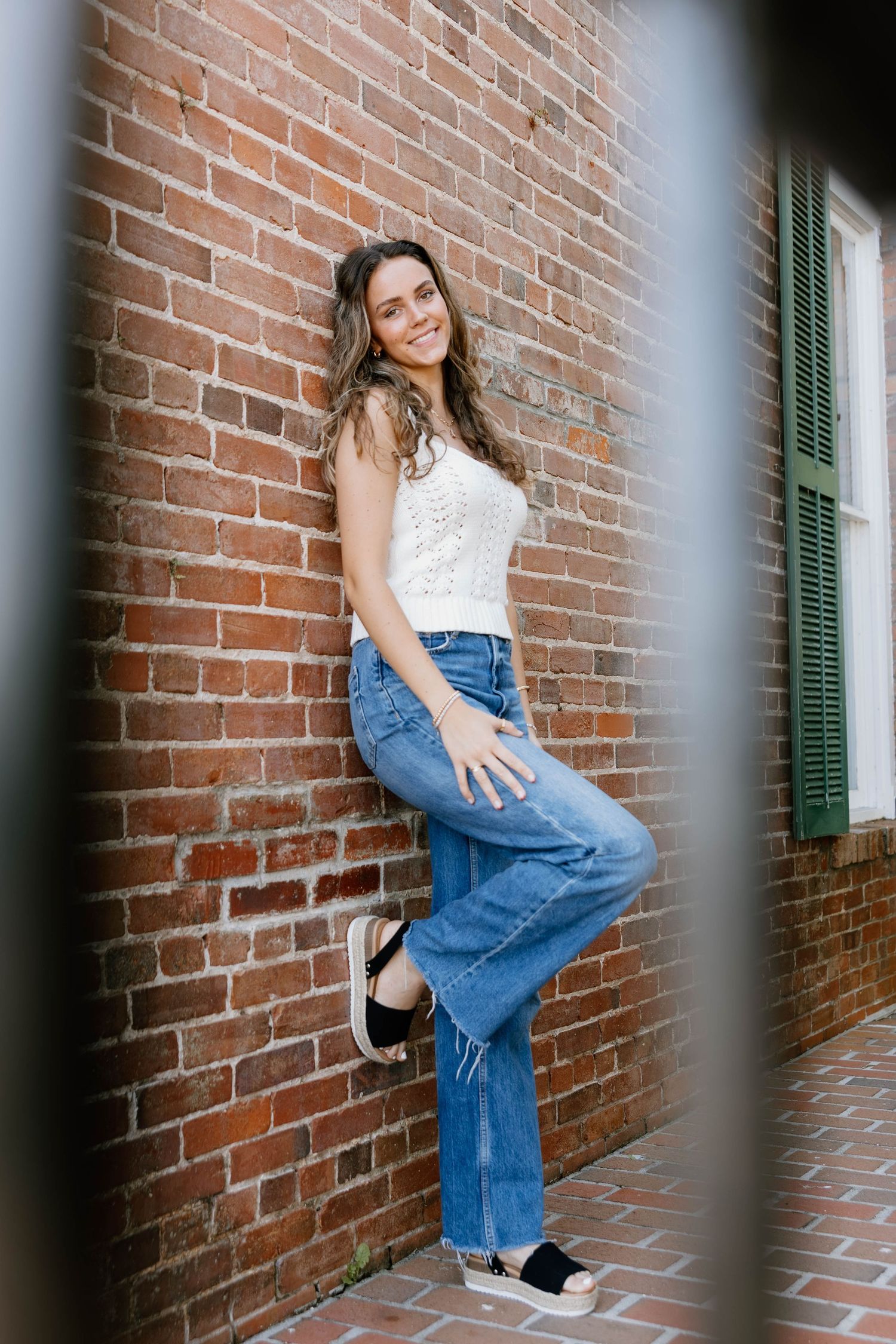 Young woman in white top and blue jeans leaning against a red brick wall, smiling outdoors.