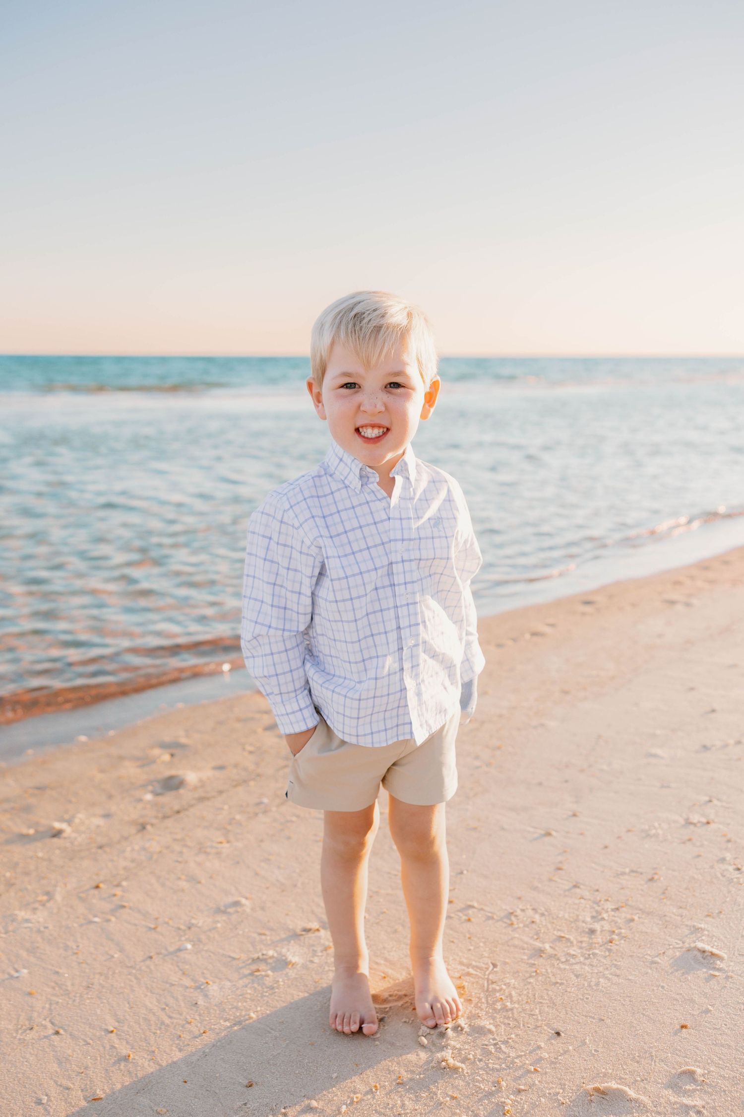 A smiling blonde boy in a white shirt and khaki shorts stands barefoot on a sandy beach with calm blue ocean waves.