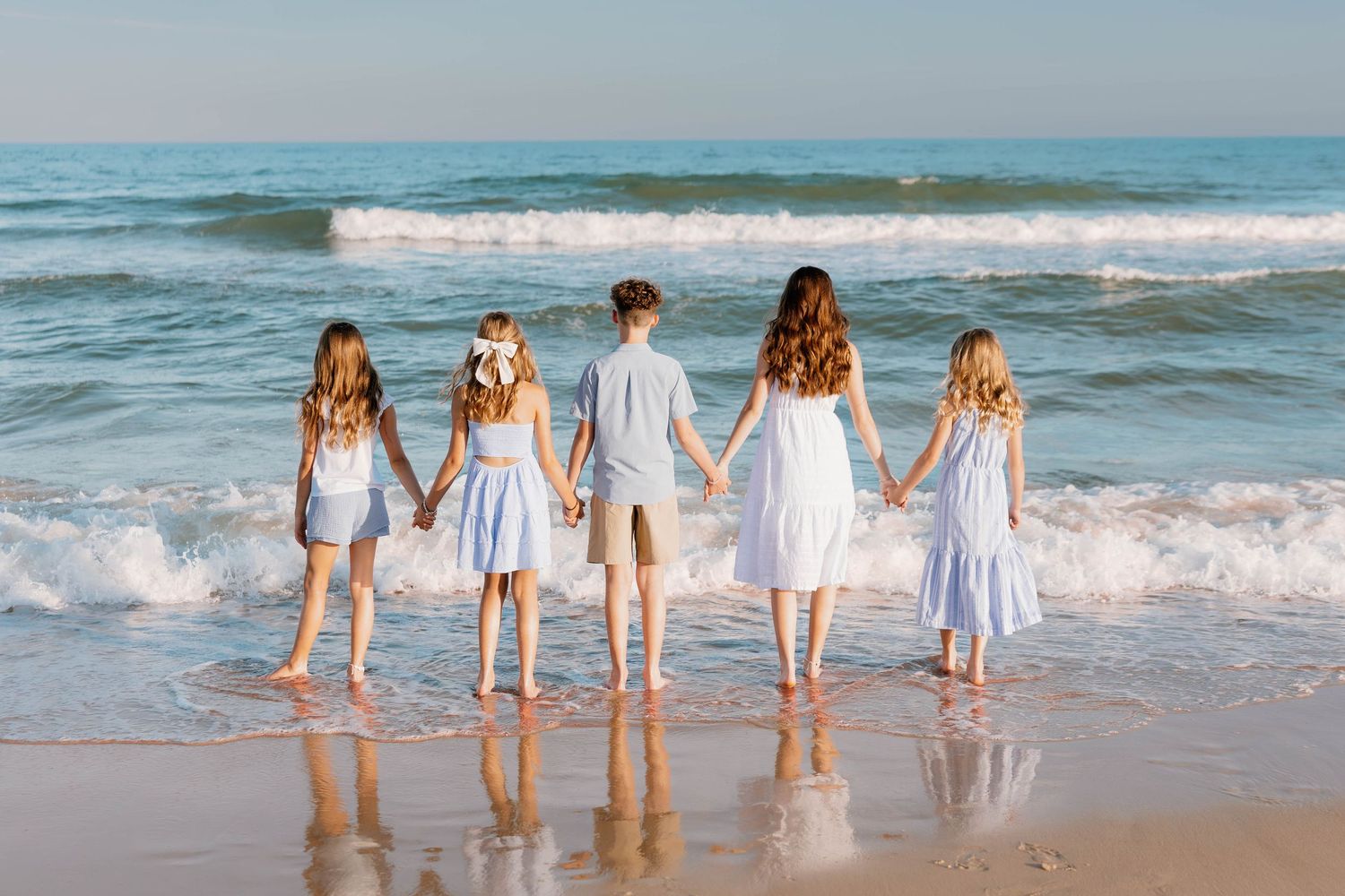 Five children in white and blue dresses hold hands facing ocean waves on a sunny beach.