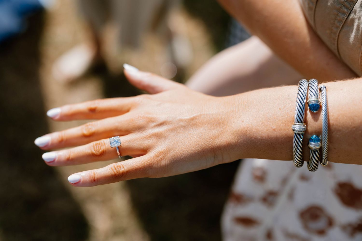Close-up of a woman's hand wearing a diamond ring and silver cable bracelets with blue gemstones outdoors.