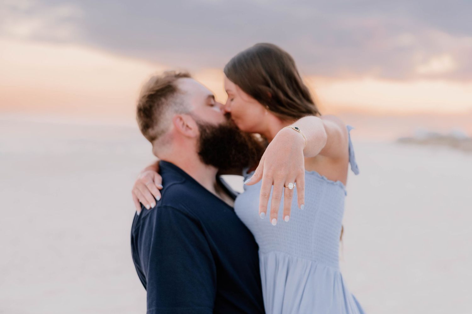 Couple sharing a romantic kiss on a sandy beach at sunset, woman wearing a light blue dress and engagement ring.