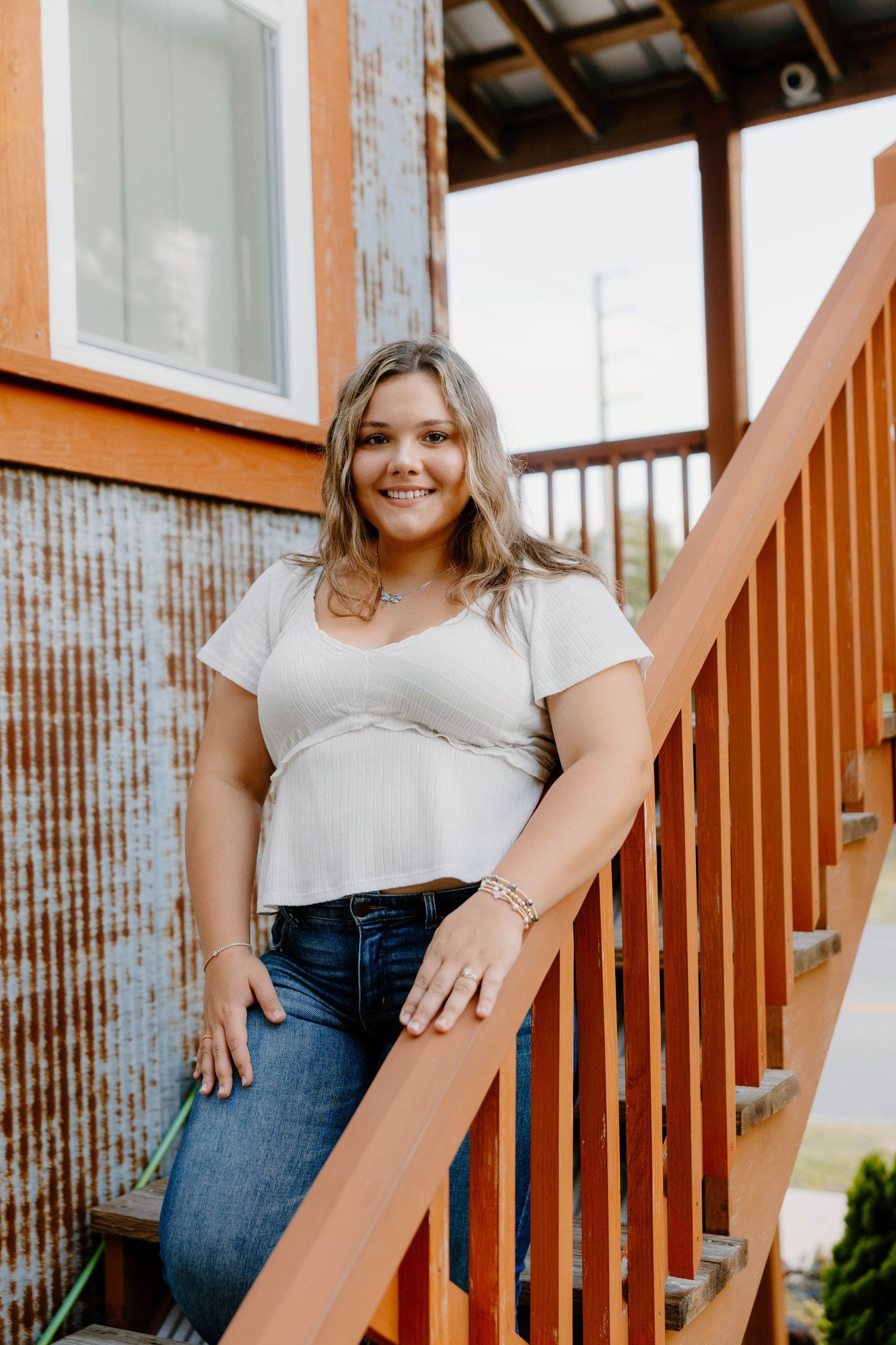 Young woman in white top and jeans smiling on wooden staircase outside rustic building.