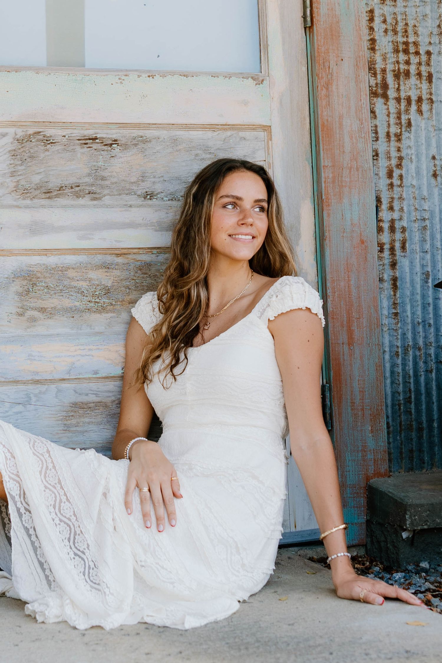 Young woman in white lace dress sitting gracefully against rustic weathered wooden wall, smiling softly.