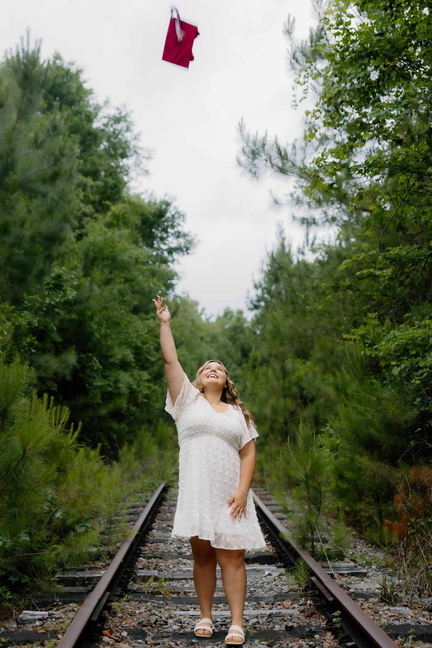 A person in a white dress poses on railroad tracks while holding a red graduation cap.