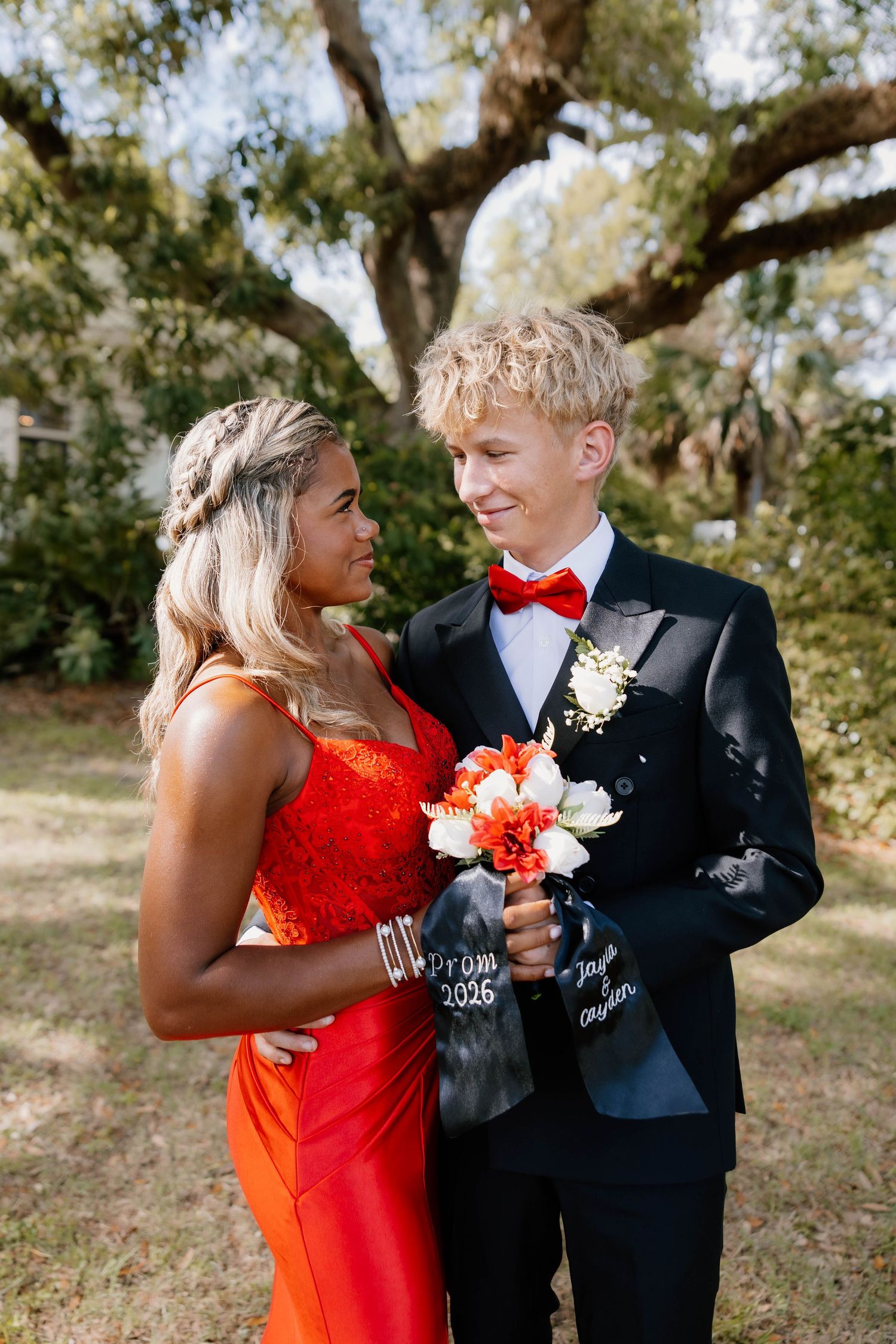 A couple dressed for prom 2026, she in red gown, he in black tuxedo with red bow tie, holding bouquet outdoors.