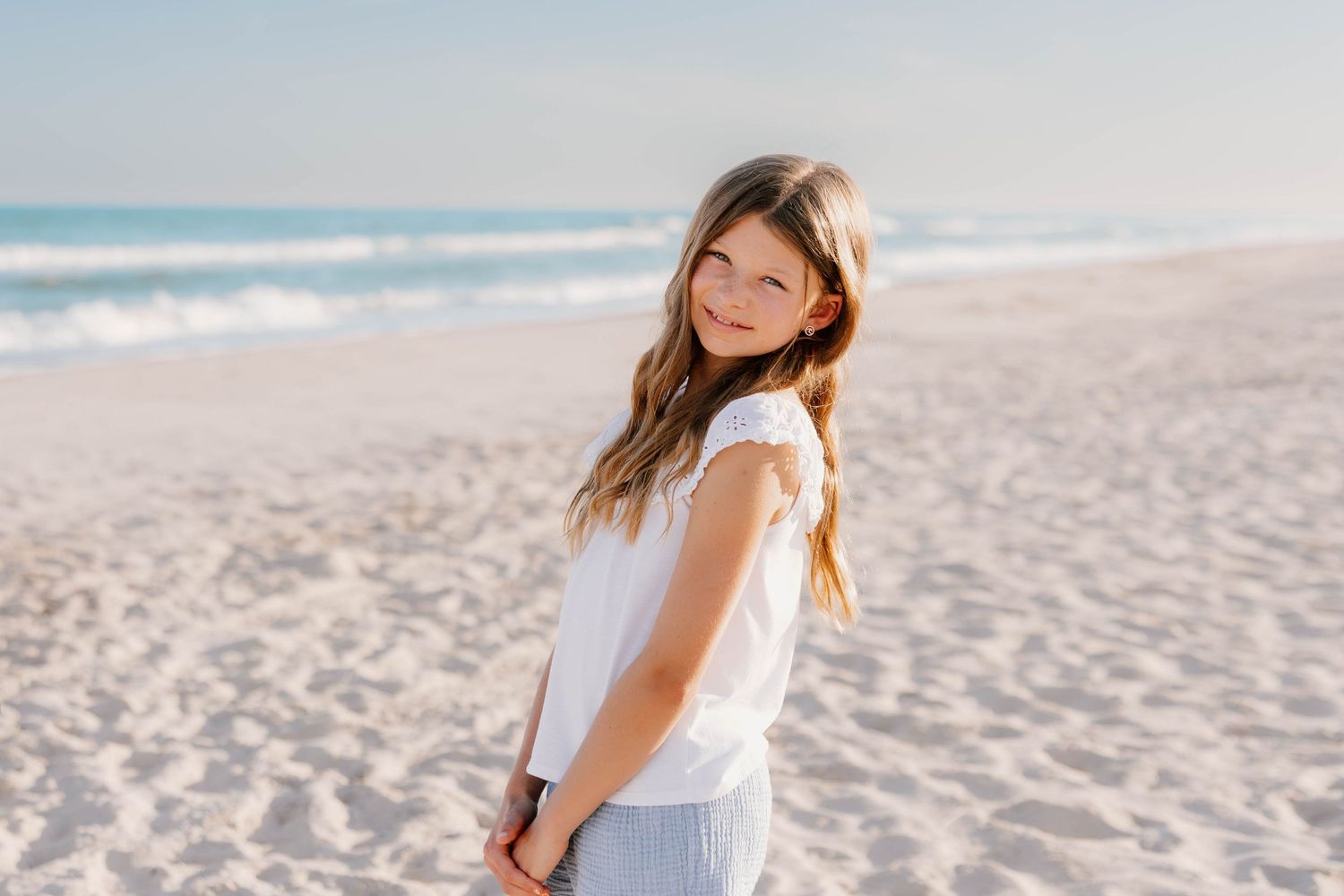 Young girl smiling over shoulder on sandy beach with ocean waves, wearing white top and blue skirt.