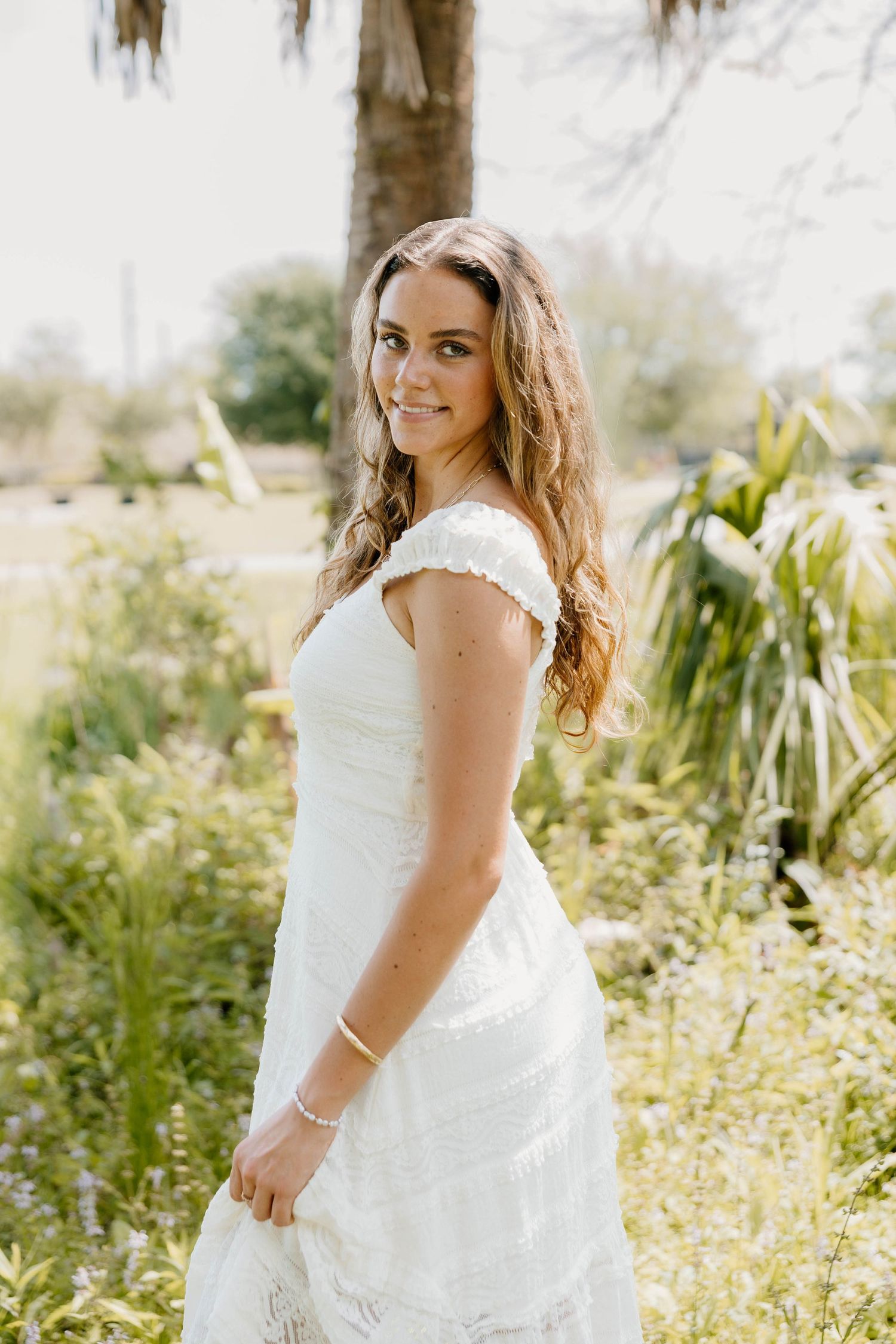 Young woman in white lace off-shoulder dress posing outdoors among lush green garden foliage.