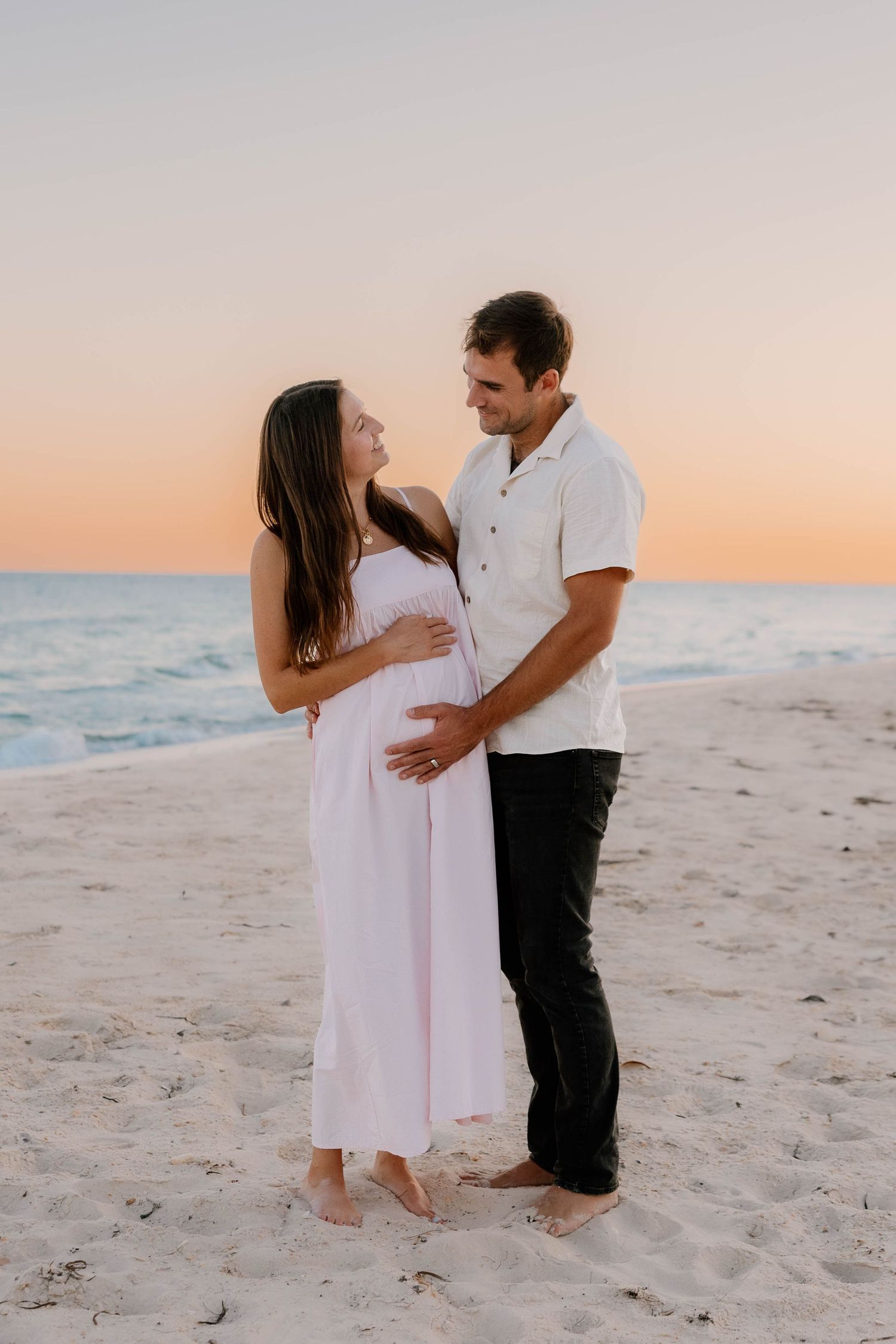 Couple embracing on a sandy beach at sunset, woman in pink maternity dress with partner in white shirt and black pants.