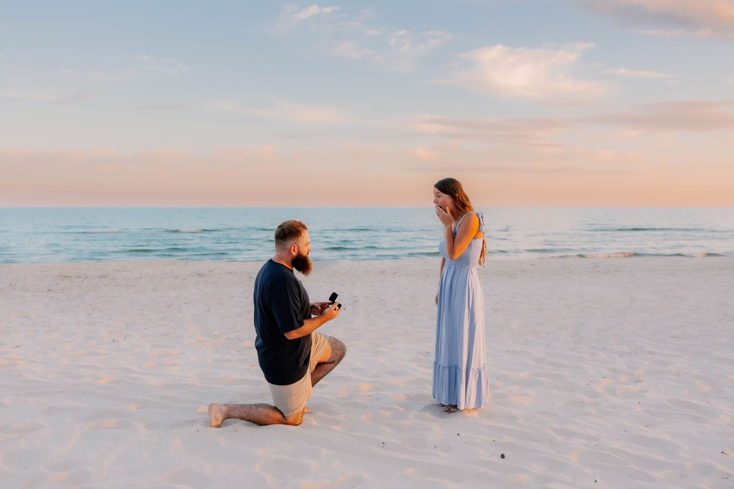 Man proposes to woman on sandy beach at sunset, kneeling with ring box as she reacts with joy.