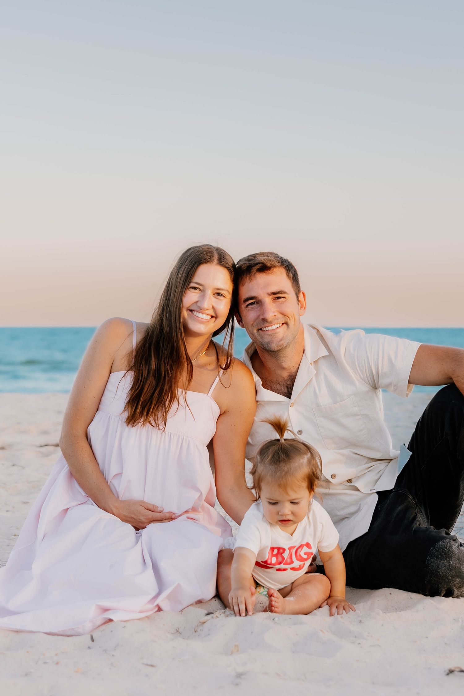 A family in white outfits poses together on a sandy beach at sunset, creating a serene coastal portrait.