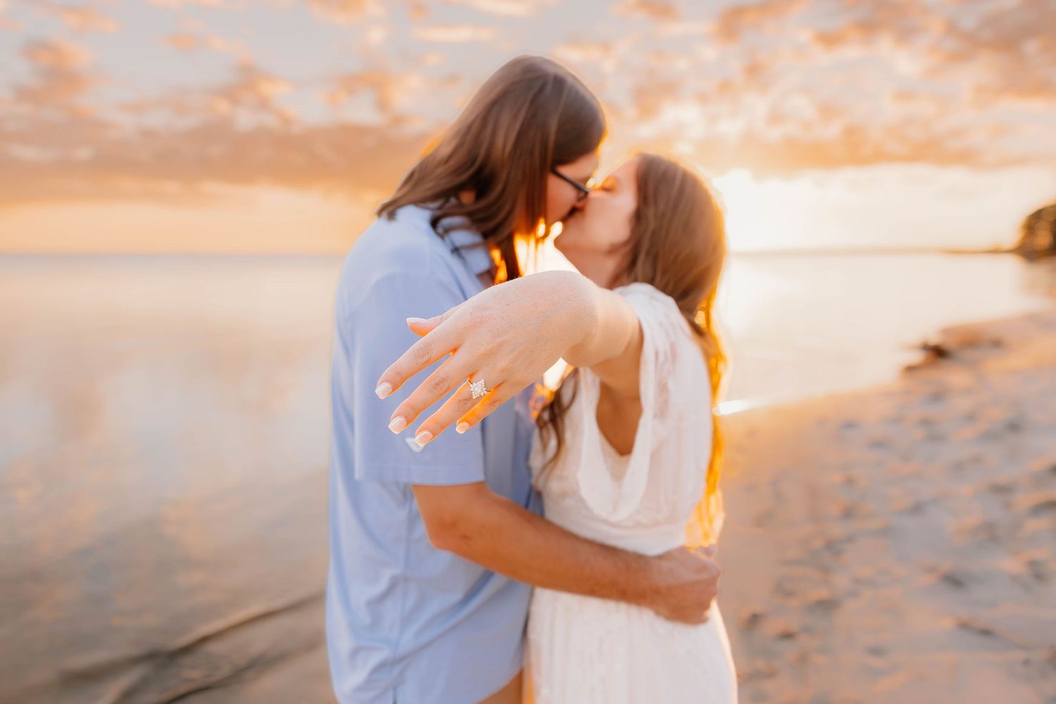Couple kissing on beach at golden sunset, woman showing engagement ring, romantic proposal moment by the water.