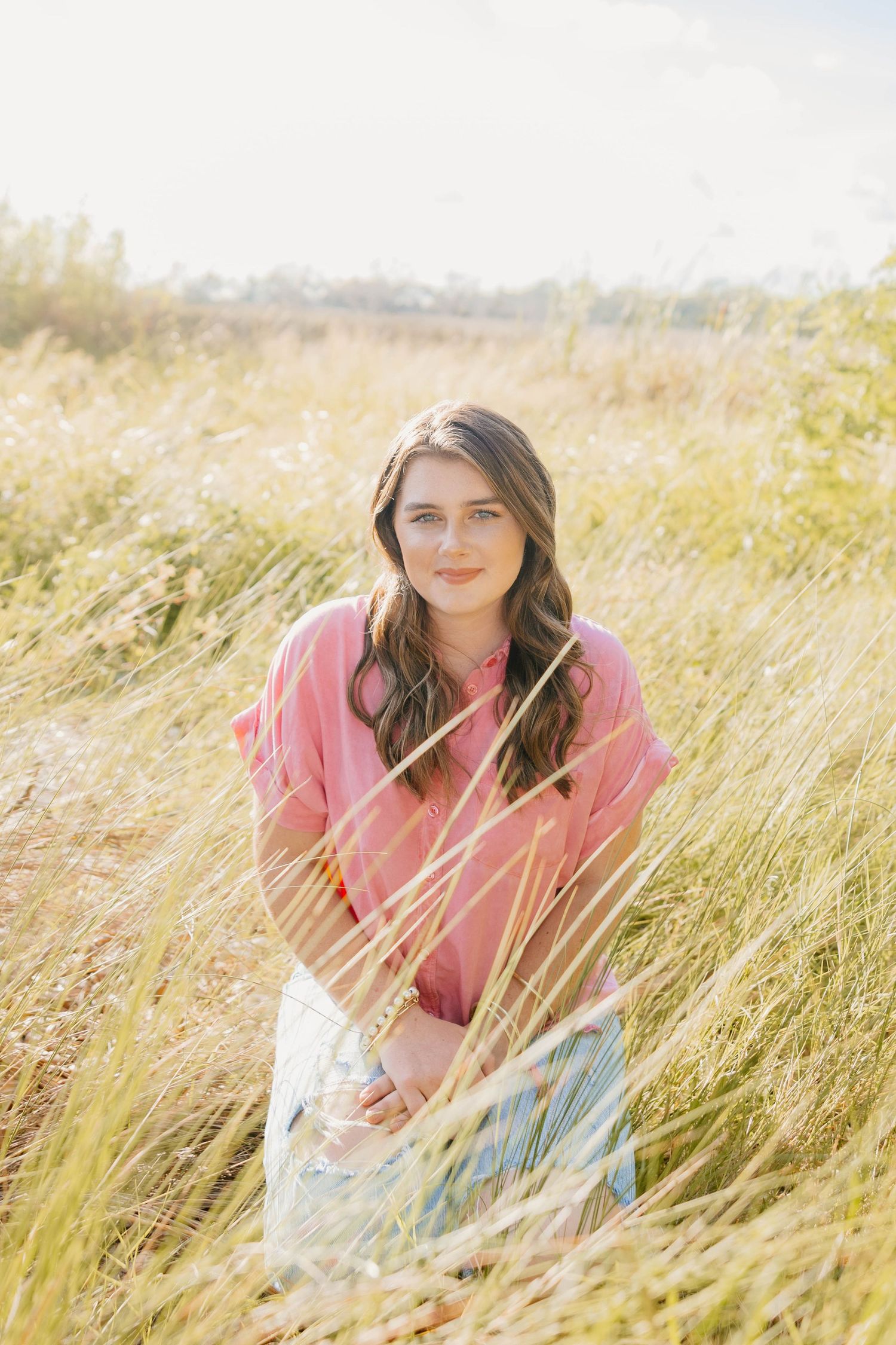 Young woman in pink top sitting in golden grass field, smiling outdoors in warm sunlight.