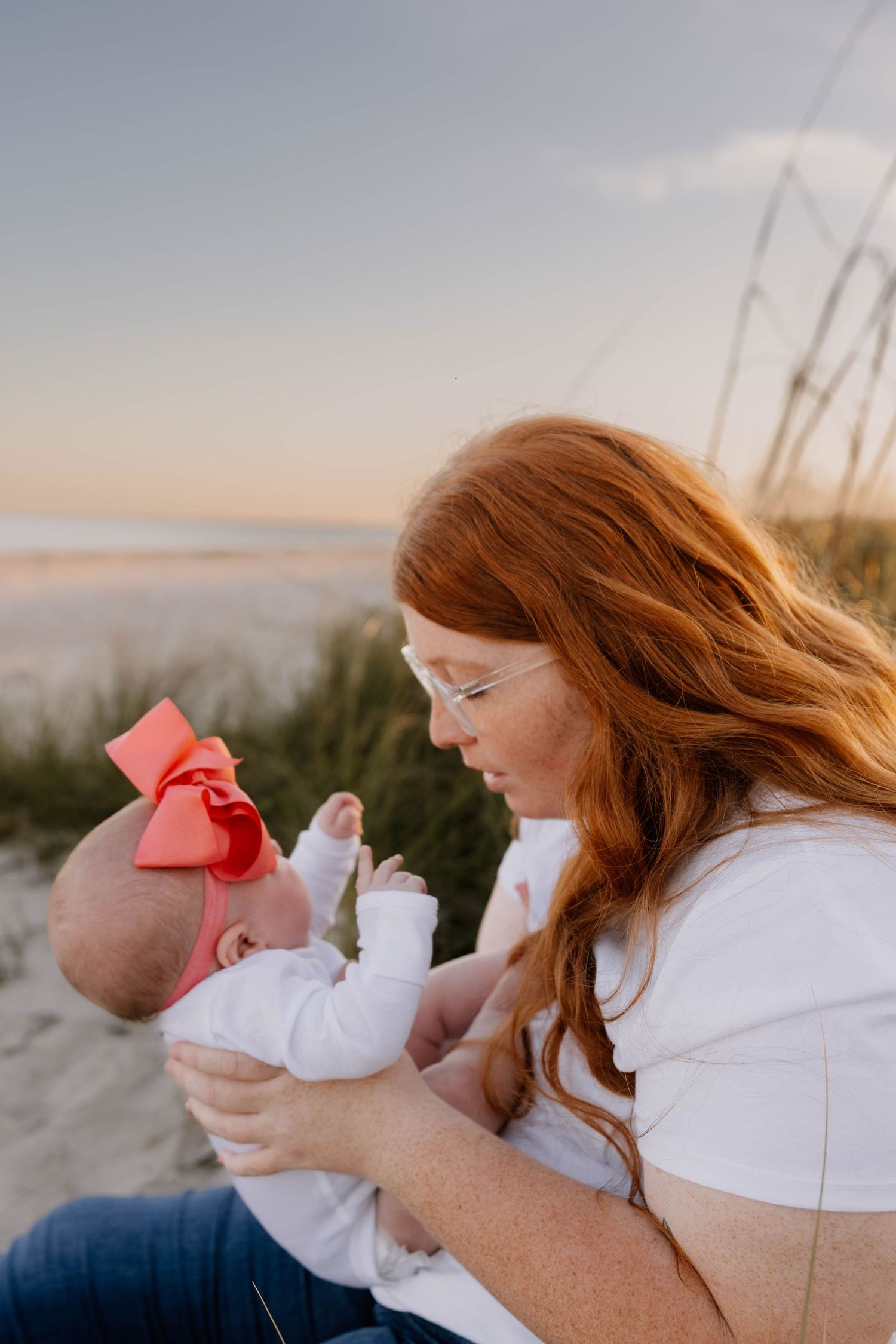 Red-haired mother tenderly holds newborn baby with pink bow on beach at sunset.
