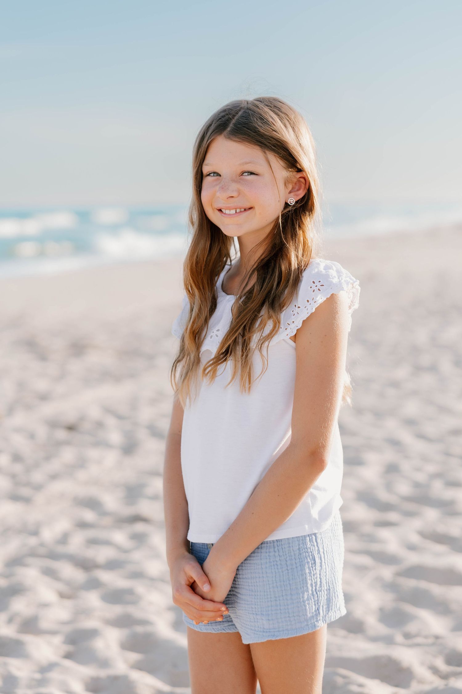 Smiling young girl with long hair wearing a white top and blue shorts standing on a sunny sandy beach.