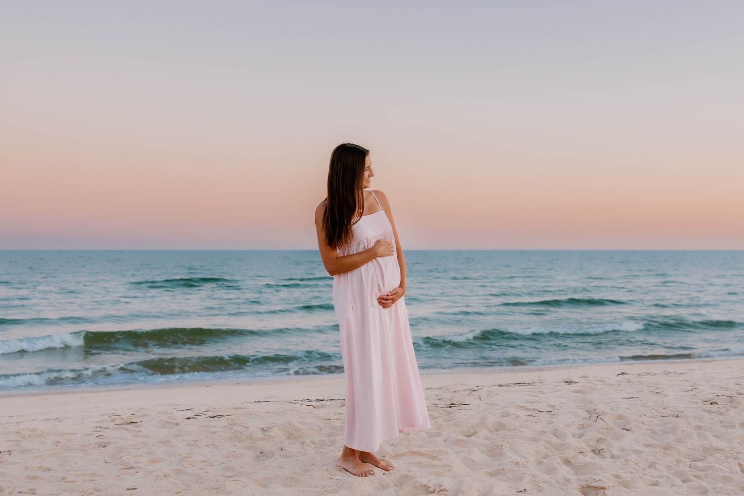 A person in a white dress walks along a serene beach at sunset with ocean waves in the background.