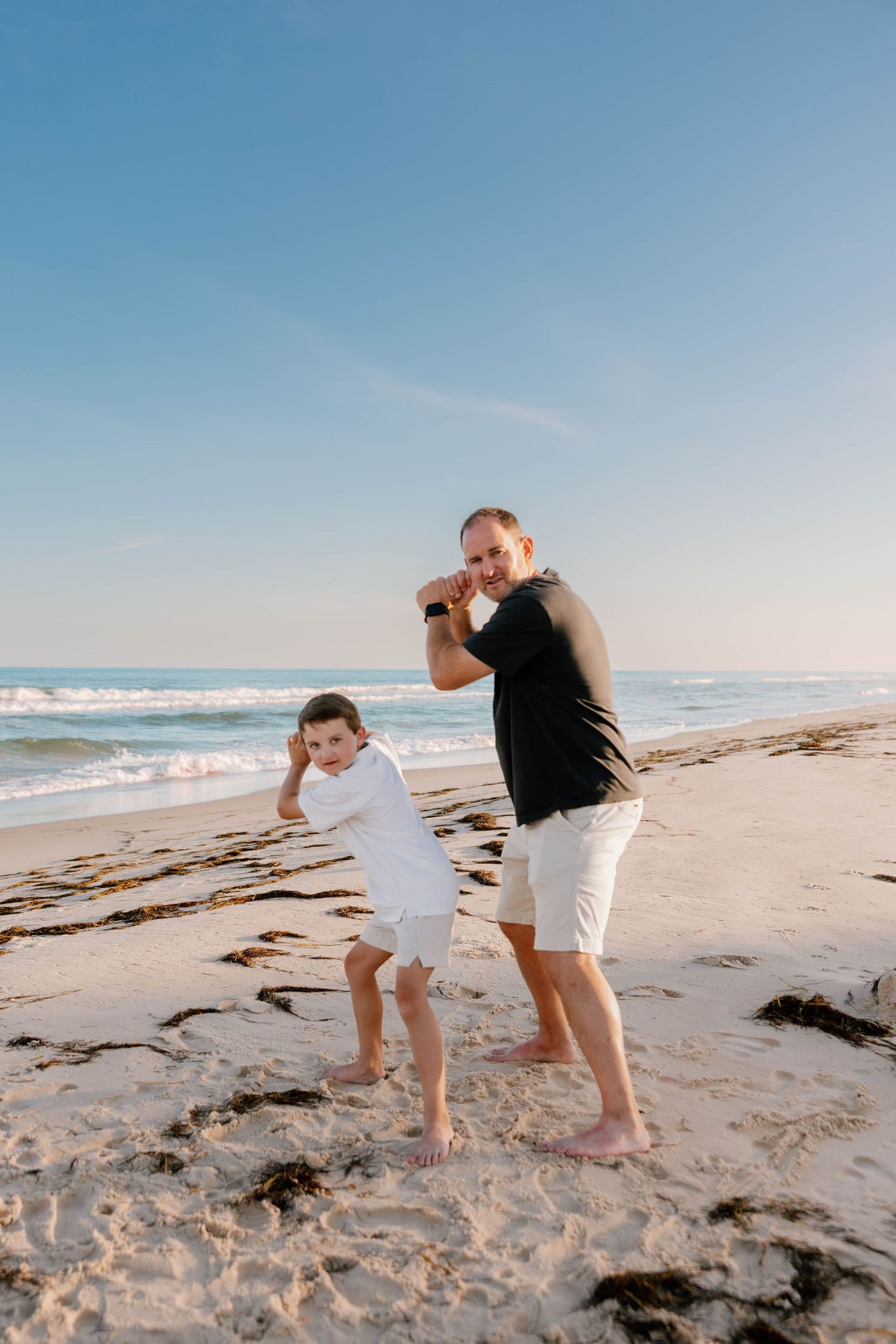 A father and son playfully boxing on a sunny sandy beach with ocean waves in the background.
